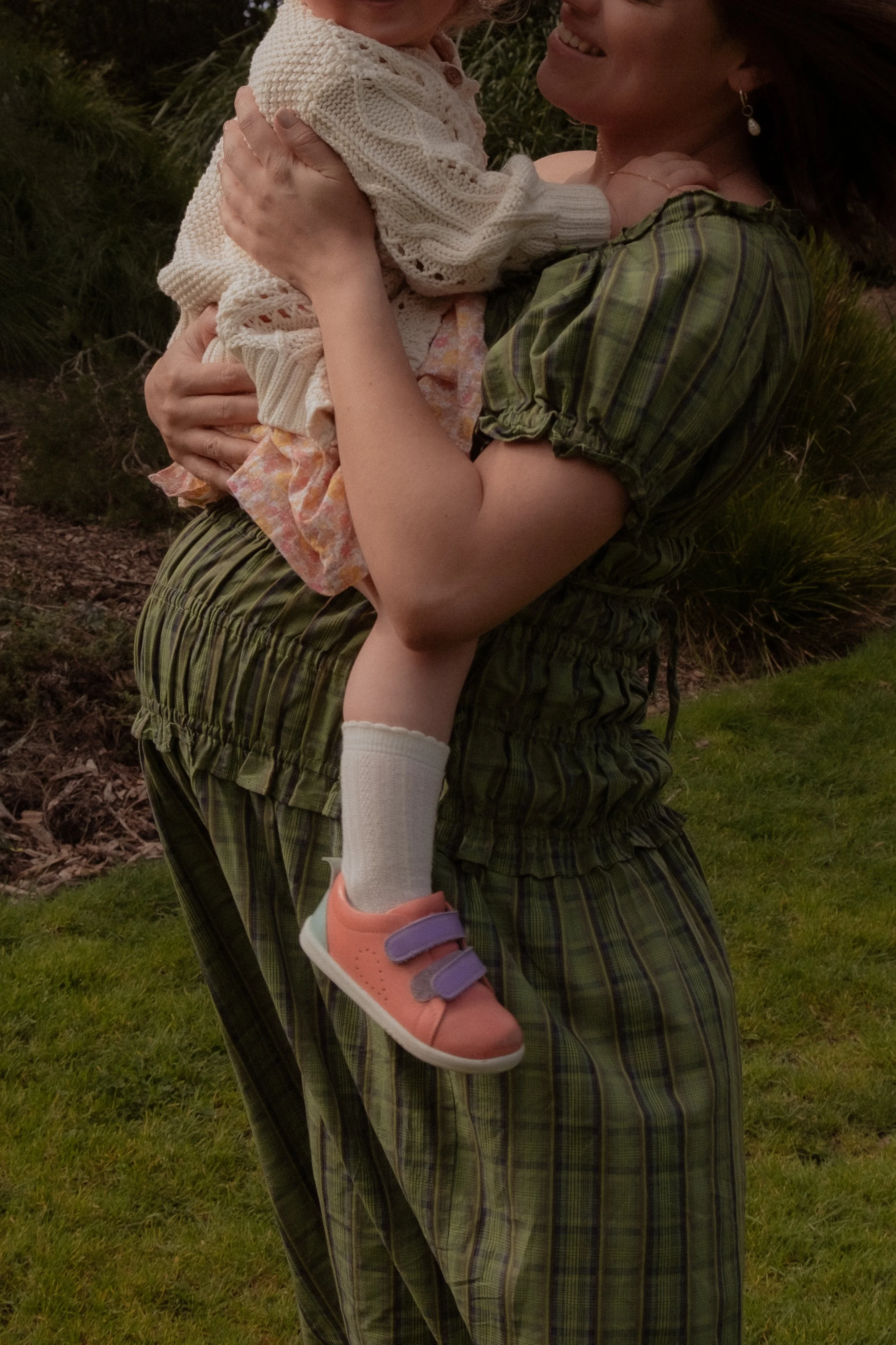A woman holding a young girl outdoors in a grassy area, both smiling. The woman is dressed in a green plaid outfit, and the girl is wearing a white sweater, colorful dress, white socks, and pink shoes with purple straps. Maternity photography. 
