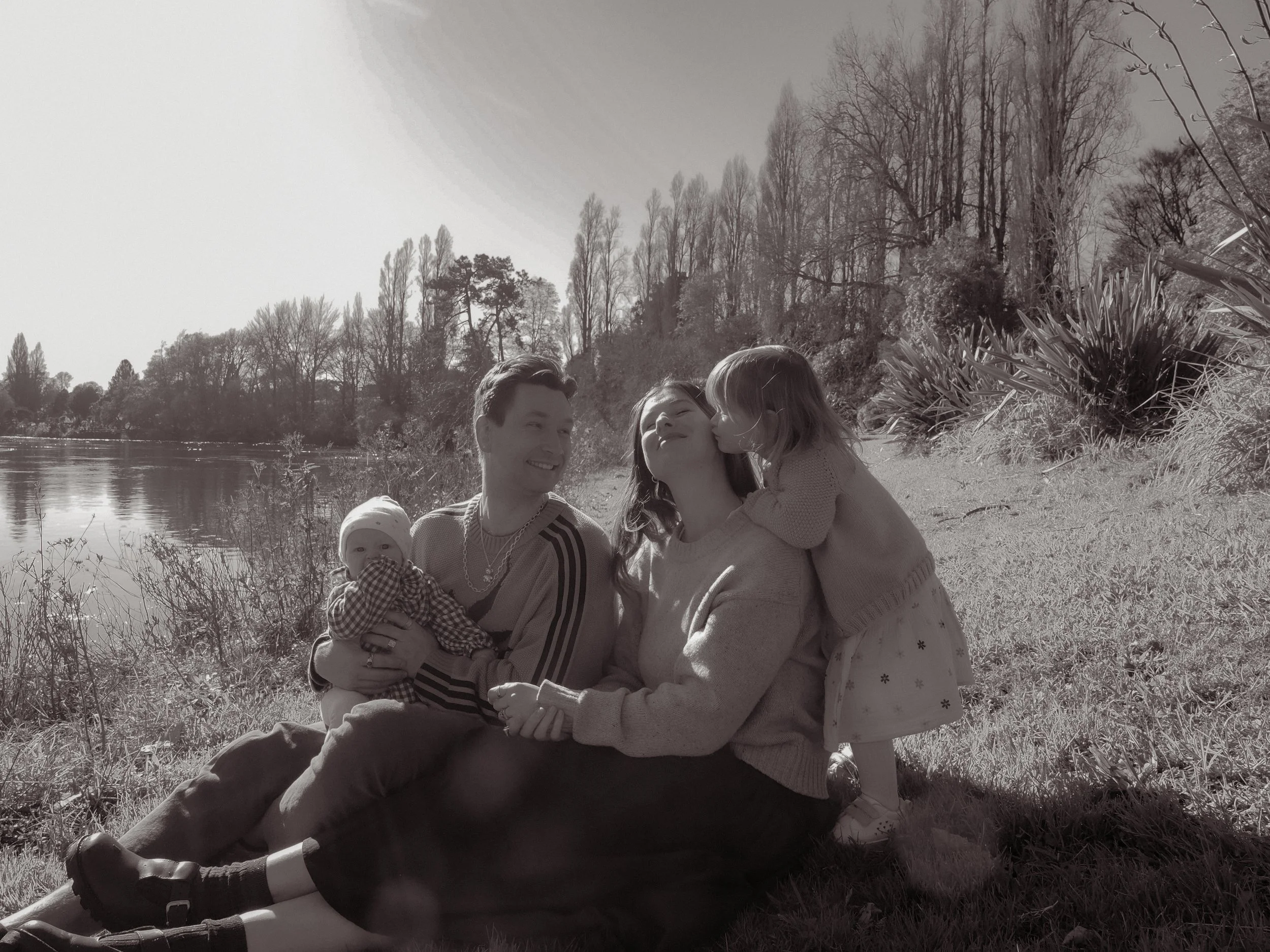 A family of four sitting on the grass beside a river, smiling and enjoying a sunny day outdoors. The parents are holding their two young children, one girl giving her mother a kiss on the cheek.