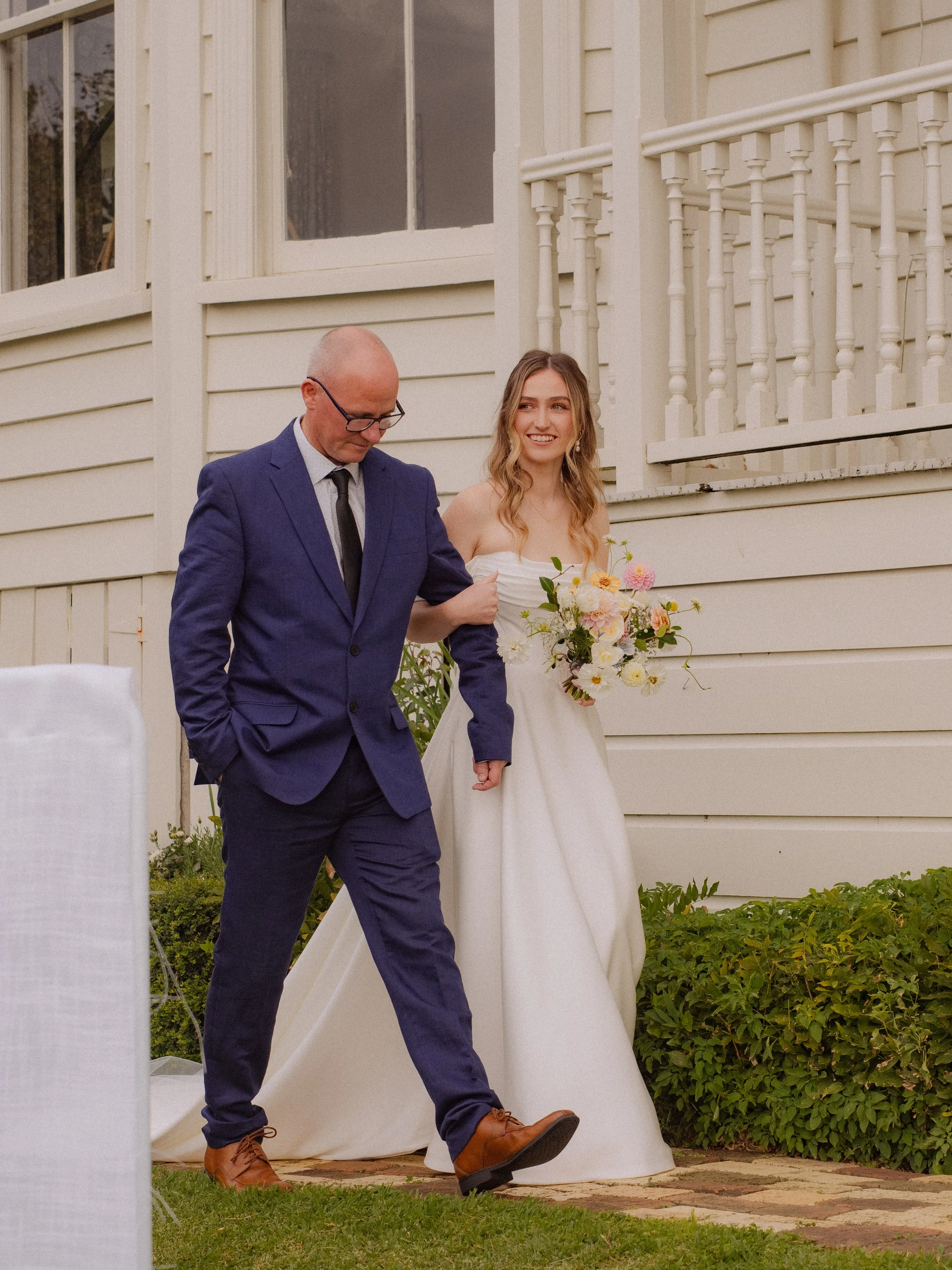 A man walking a woman down the aisle at an outdoor wedding ceremony, with the woman holding a bouquet of flowers and wearing a white wedding dress.  Wedding Photography Waikato New Zealand. Wedding Photography Hamilton New Zealand. Northland Wedding 