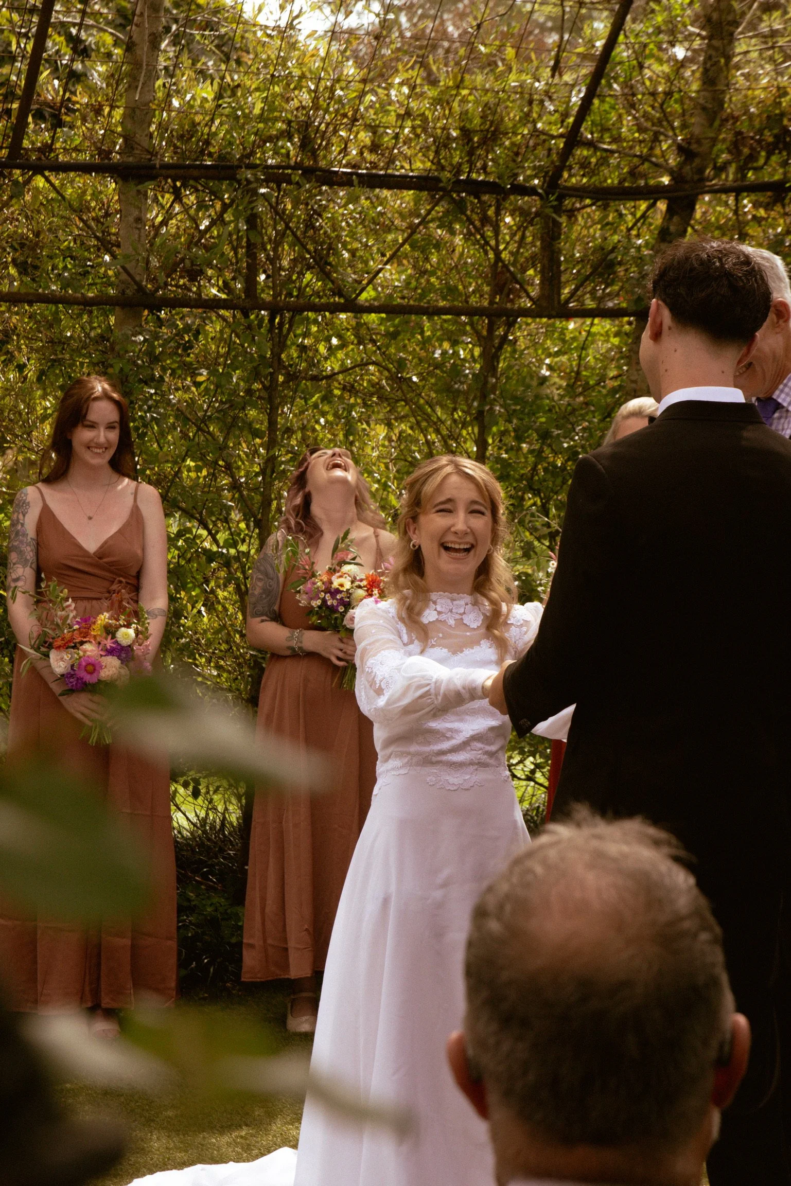 A bride and groom exchanging vows during a wedding ceremony outdoors in a wooded area, with bridesmaids standing behind them holding bouquets and smiling. Wedding Photography Waikato New Zealand. Wedding Photography Hamilton New Zealand