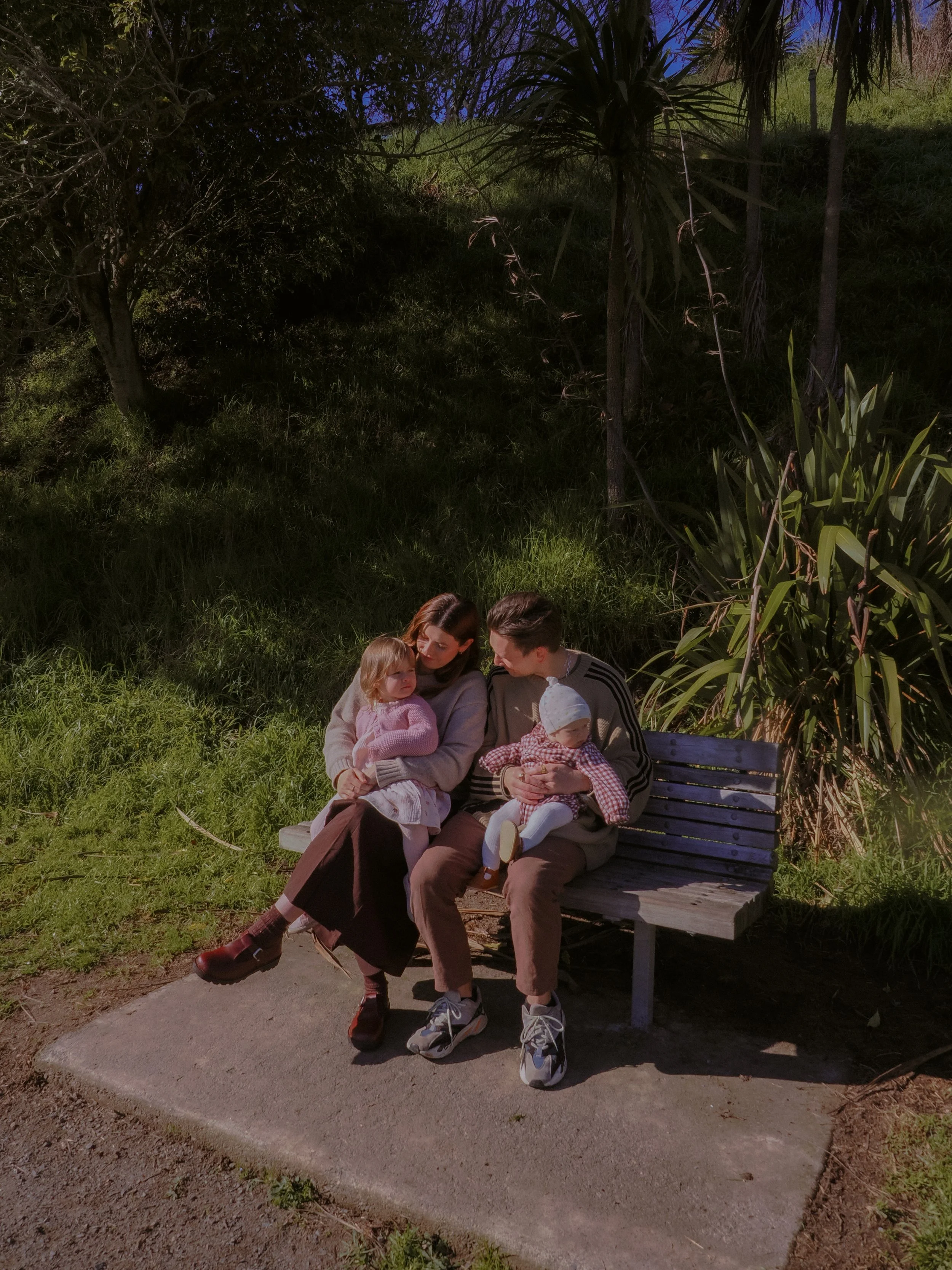 Family of four sitting on a park bench at night, surrounded by greenery, with a mother, father, and two young children. Family Photography Waikato New Zealand. Family Photography Hamilton New Zealand. Northland Wedding Photography