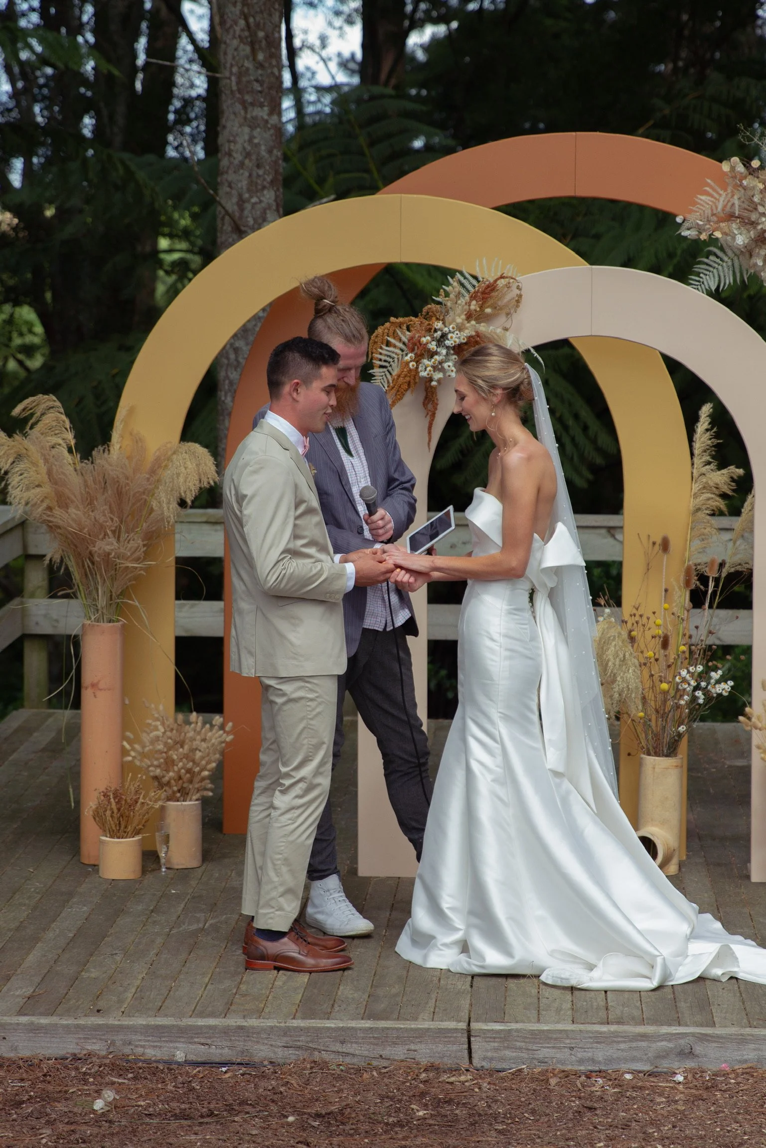 A wedding ceremony outdoors with a couple exchanging vows, an officiant holding a microphone, and floral decor in earthy tones.  Wedding Photography Waikato New Zealand. Wedding Photography Hamilton New Zealand. Northland Wedding Photography