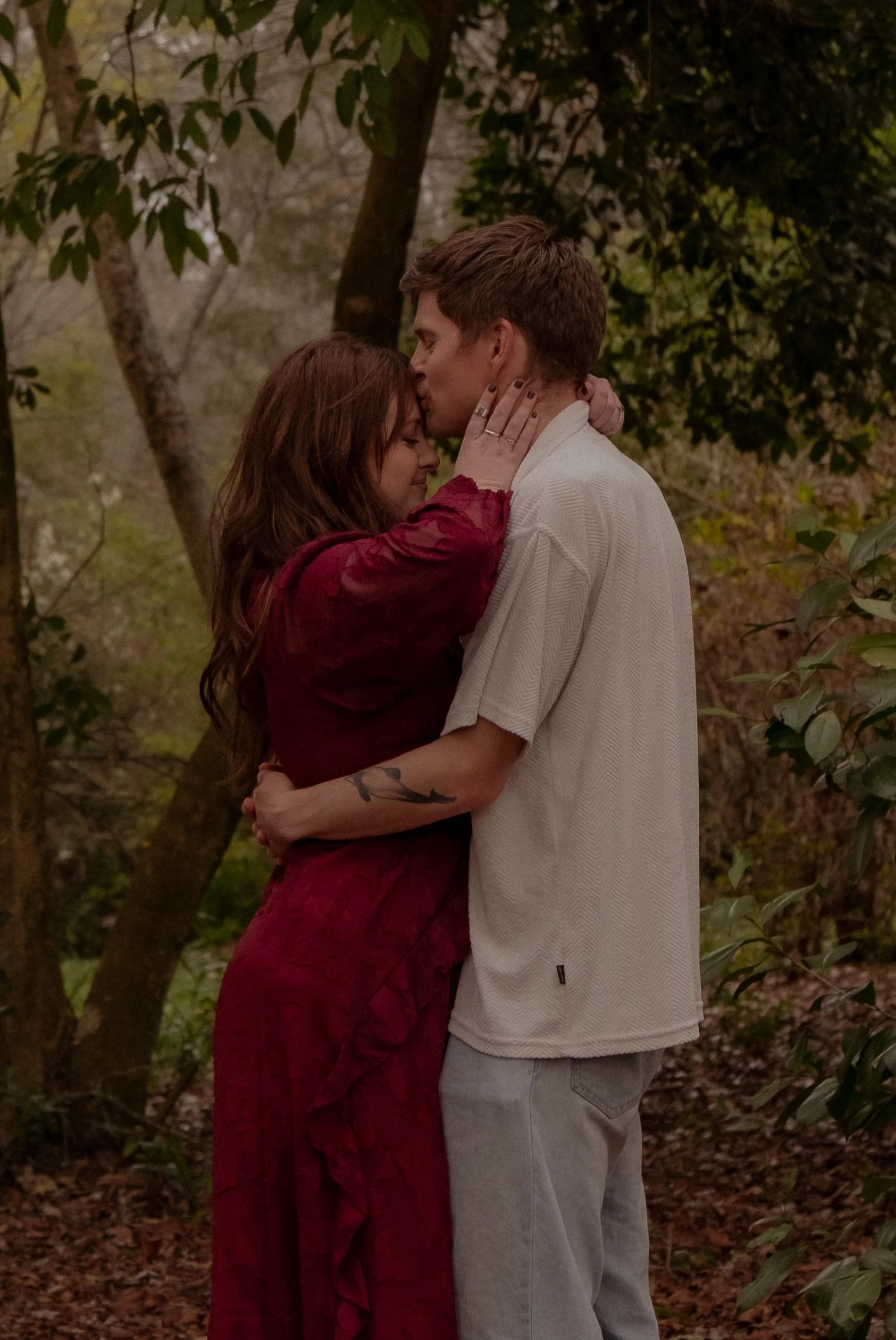 A couple embracing outdoors surrounded by trees, with the man giving the woman a kiss on the forehead. Couples photography Hamilton New Zealand. Engagement photography Hamilton New Zealand. Whangarei New Zealand Couples photographer