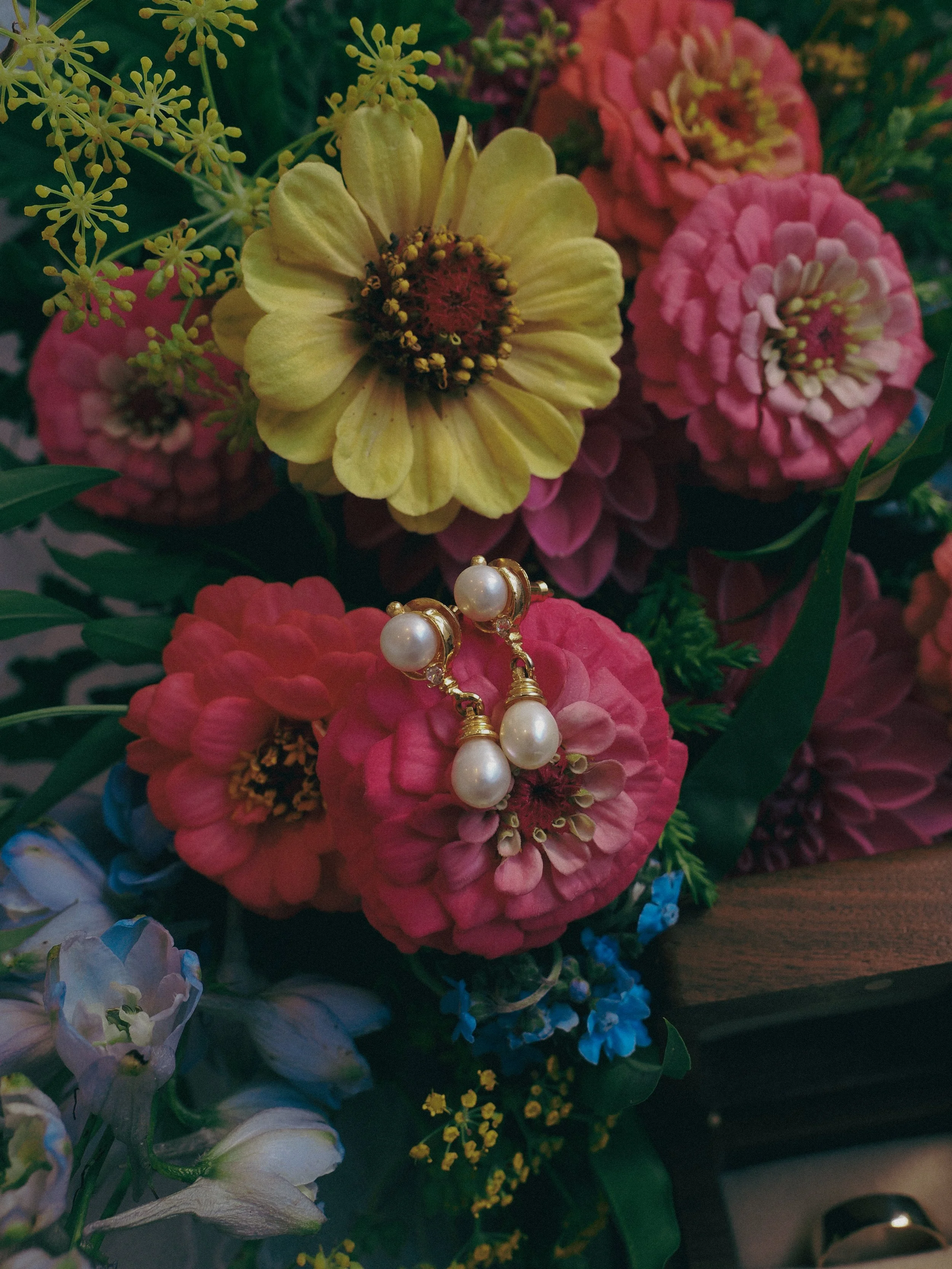 Close-up of a bouquet of mixed flowers, including pink, yellow, and white blooms, with a pair of pearl earrings resting among the flowers.