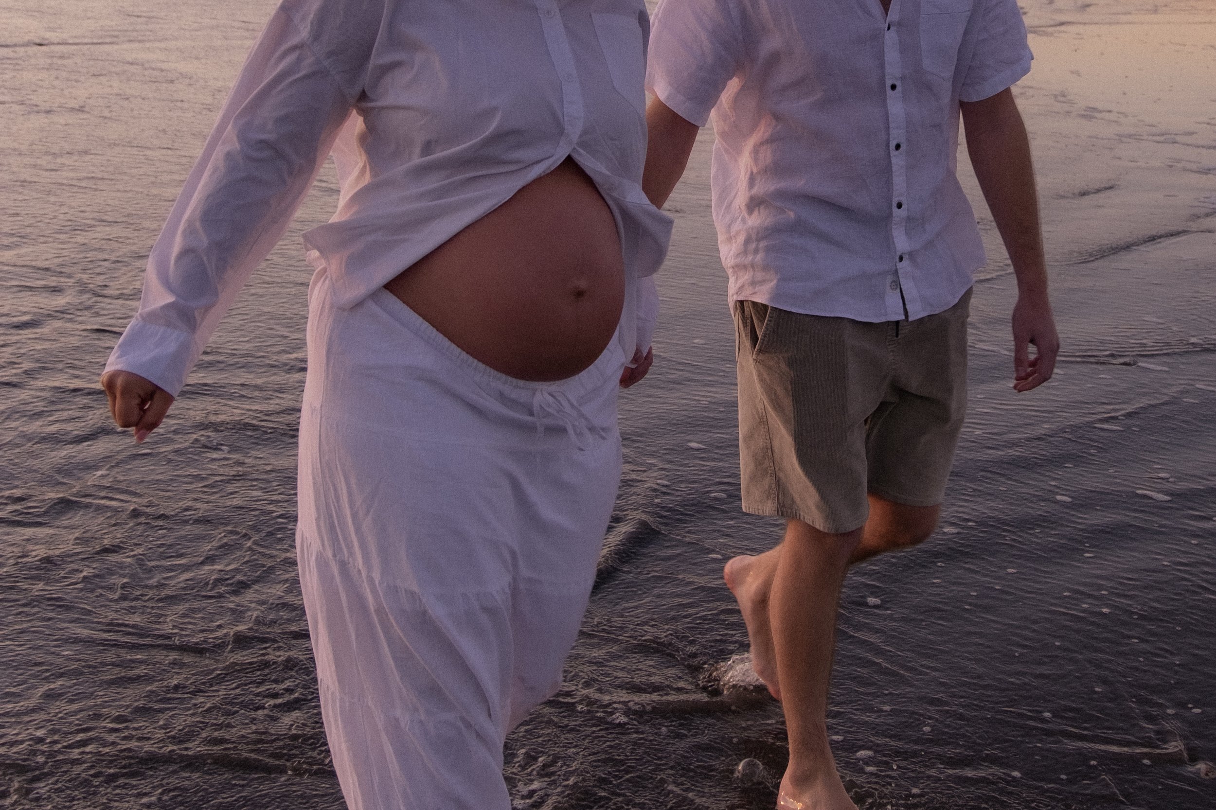 Two men in light-colored clothing walking barefoot in shallow ocean water at sunset, one with a pregnant belly. Maternity Photography. Raglan New Zealand Photographer