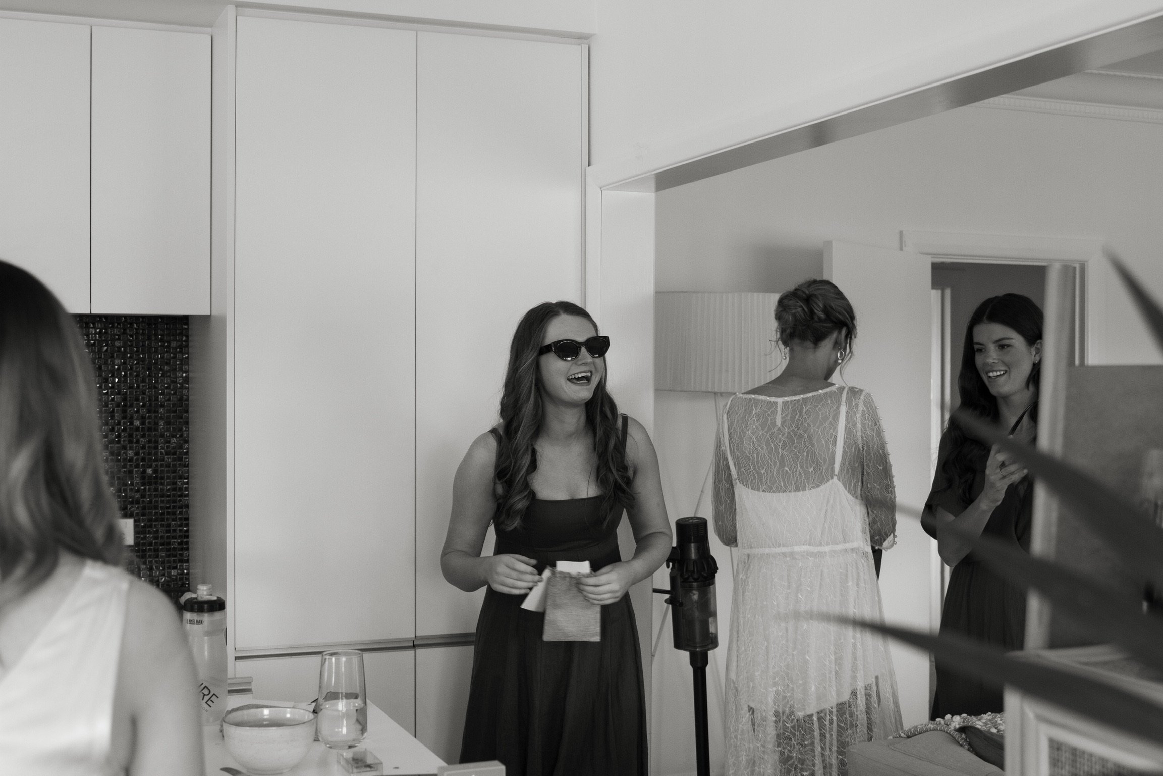 Four women in a room, with three facing the camera and smiling, one facing away. One woman wearing sunglasses and a black dress is holding a tissue and smiling, while the other women are talking and smiling.