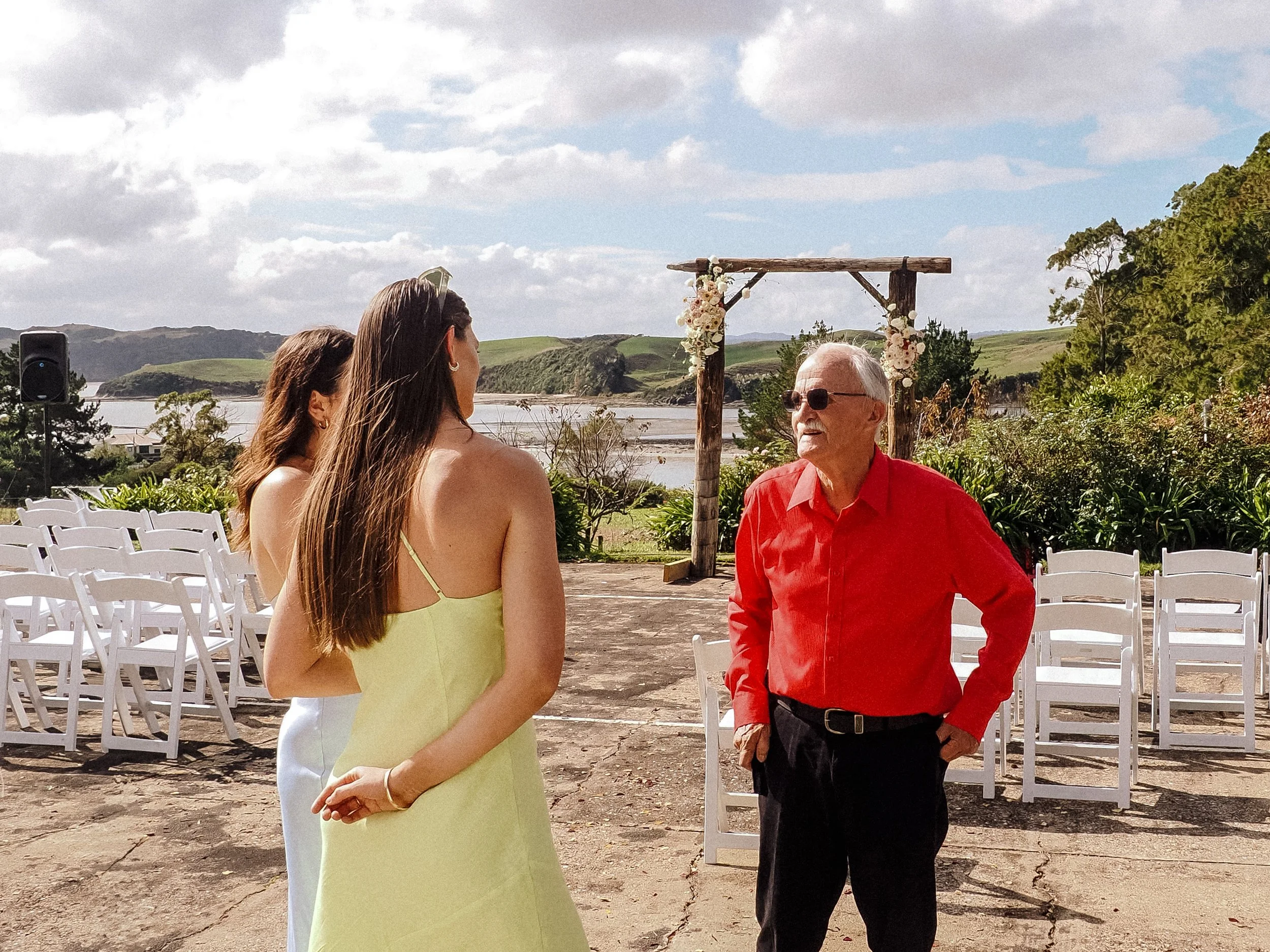 Three people engaged in conversation at an outdoor wedding ceremony site by a body of water with green hills in the background. An arch decorated with flowers is behind them, and rows of white chairs are set for guests.