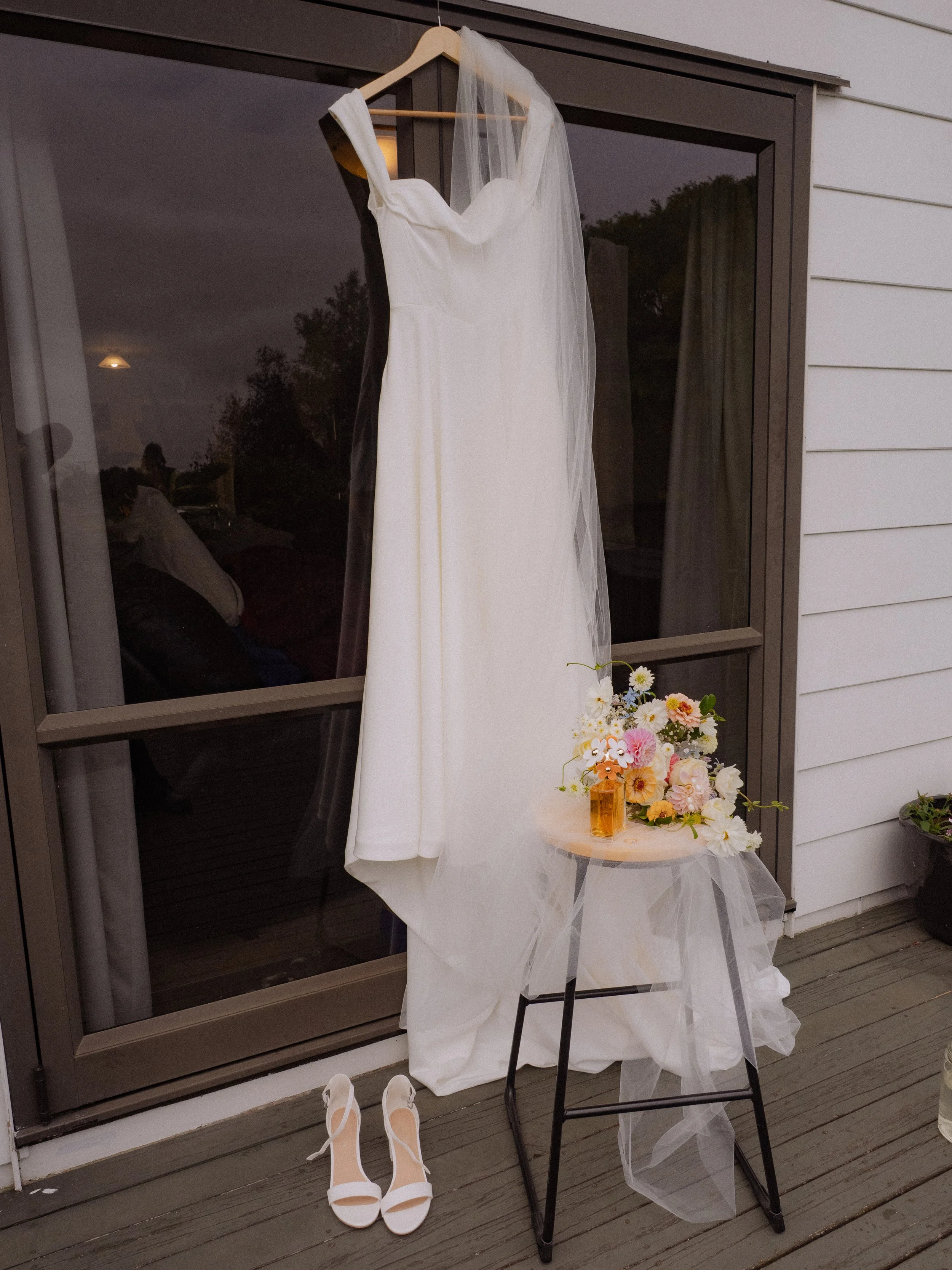 A white wedding dress and veil hang on a hanger outside a building. A pair of white high-heeled shoes sit on the ground below. A small table next to the dress holds a bouquet of pink and white flowers with green leaves, and a small amber glass bottle