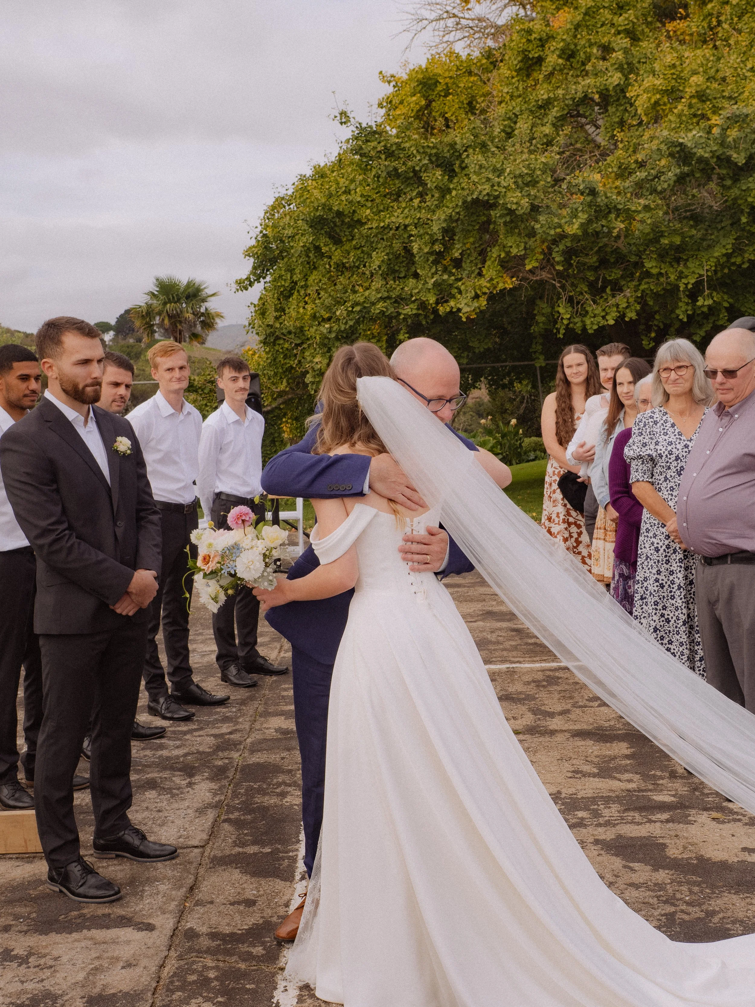 A bride and groom share a hug during their outdoor wedding ceremony, surrounded by friends and family on a cloudy day, with lush green trees in the background.  Wedding Photography Waikato New Zealand. Wedding Photography Hamilton New Zealand. 