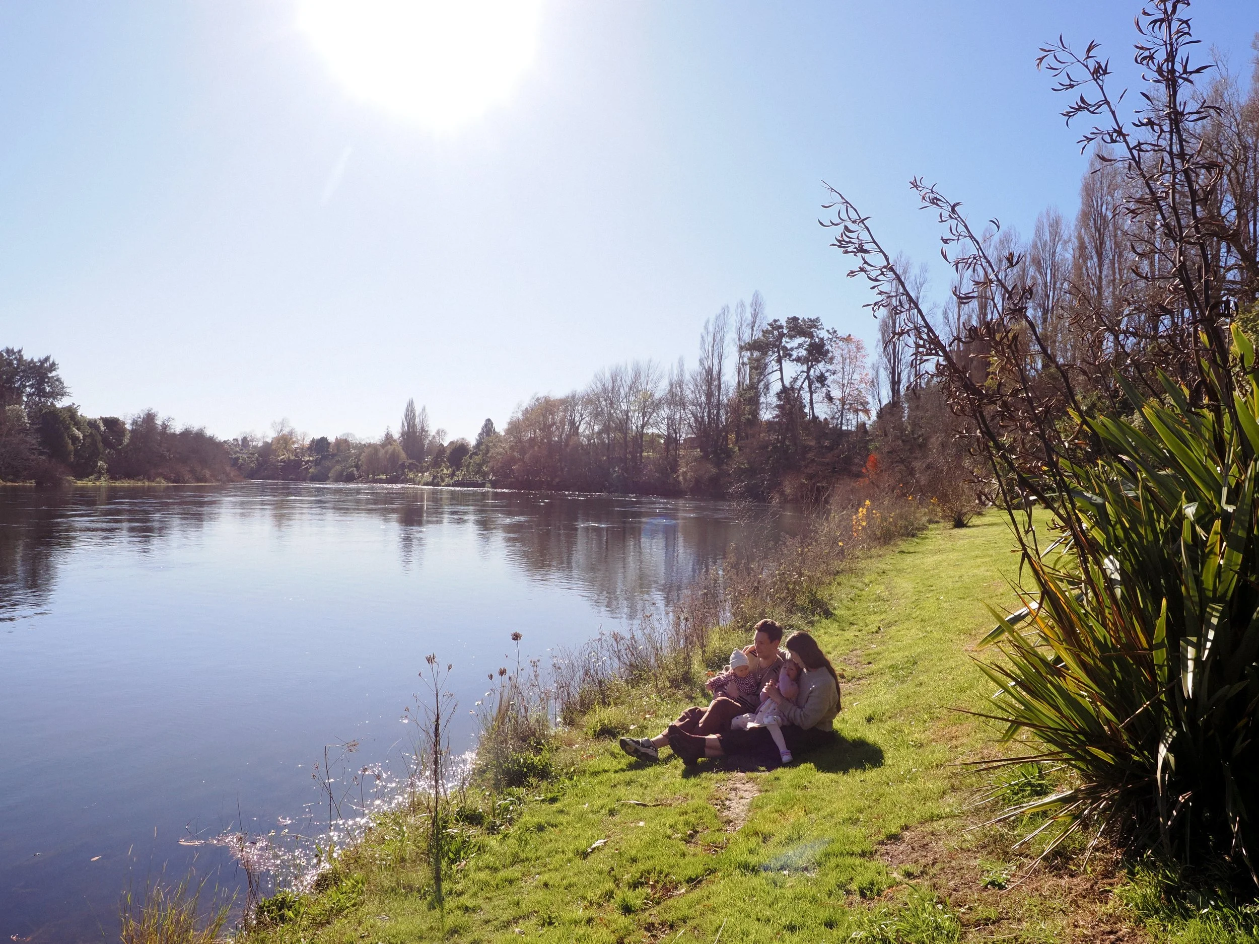 Family sitting on grassy riverbank on a sunny day, with trees and shrubs in the background, next to a calm river. Family Photography Waikato New Zealand. Family Photography Hamilton New Zealand. Northland Wedding Photography