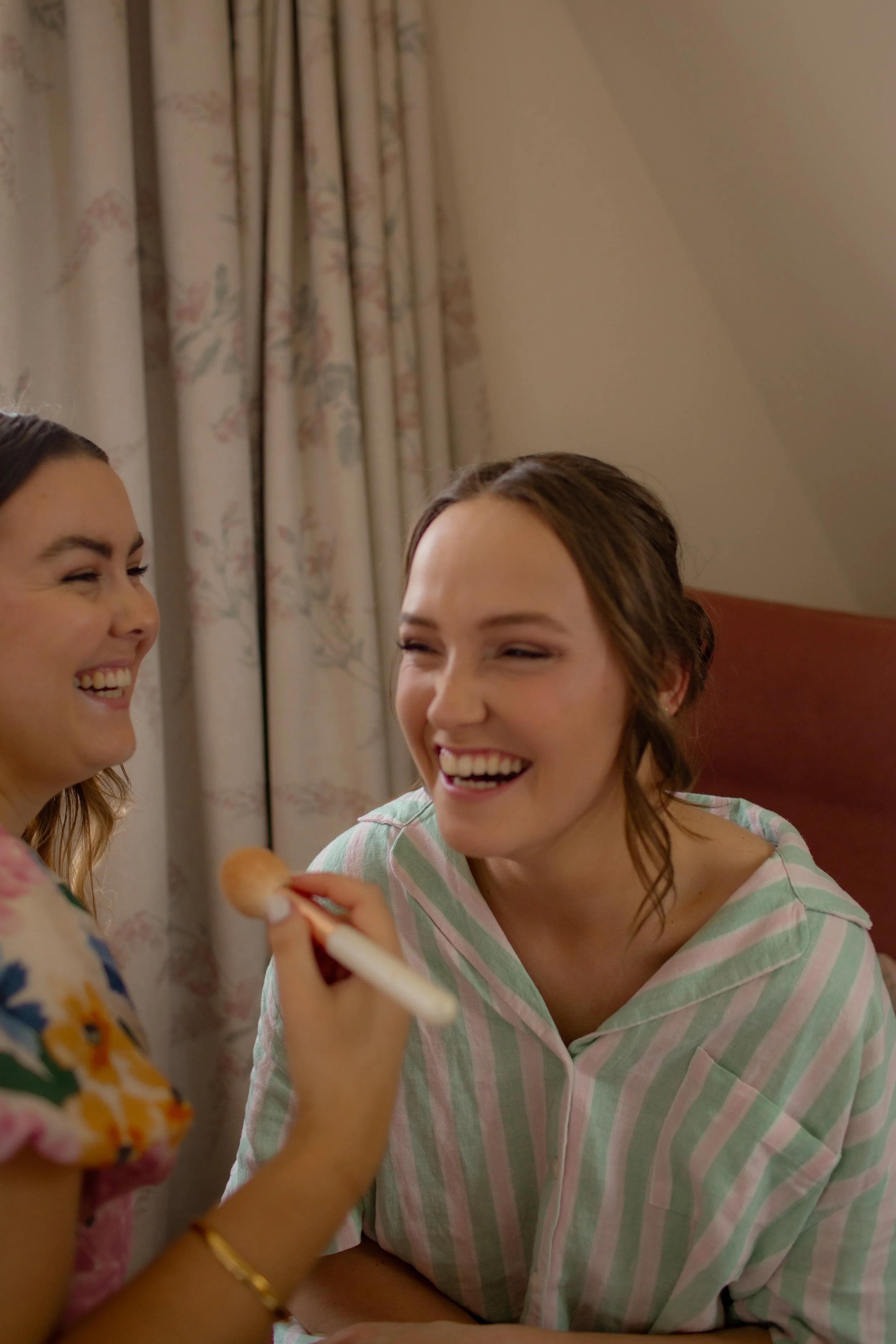 Two women smiling, one applying makeup with a brush in front of a curtain in a room with angled ceiling.  Wedding Photography Waikato New Zealand. Wedding Photography Hamilton New Zealand. Northland Wedding Photography