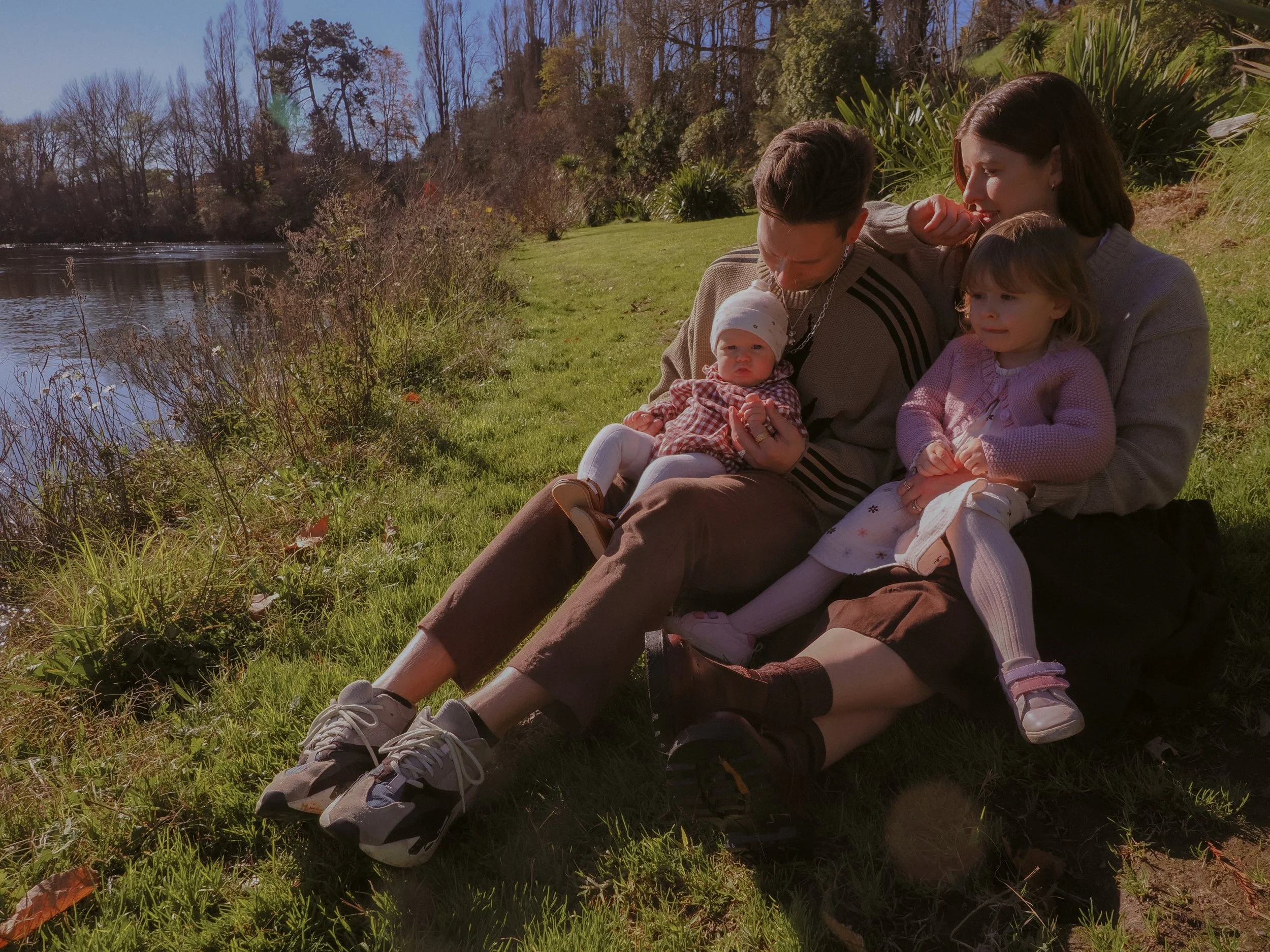 A family of four sitting on the grass by a lake in a park, with trees in the background. The father holds a baby, the mother and an older girl sit beside them. Family Photography Waikato New Zealand. Family Photography Hamilton New Zealand. Northland