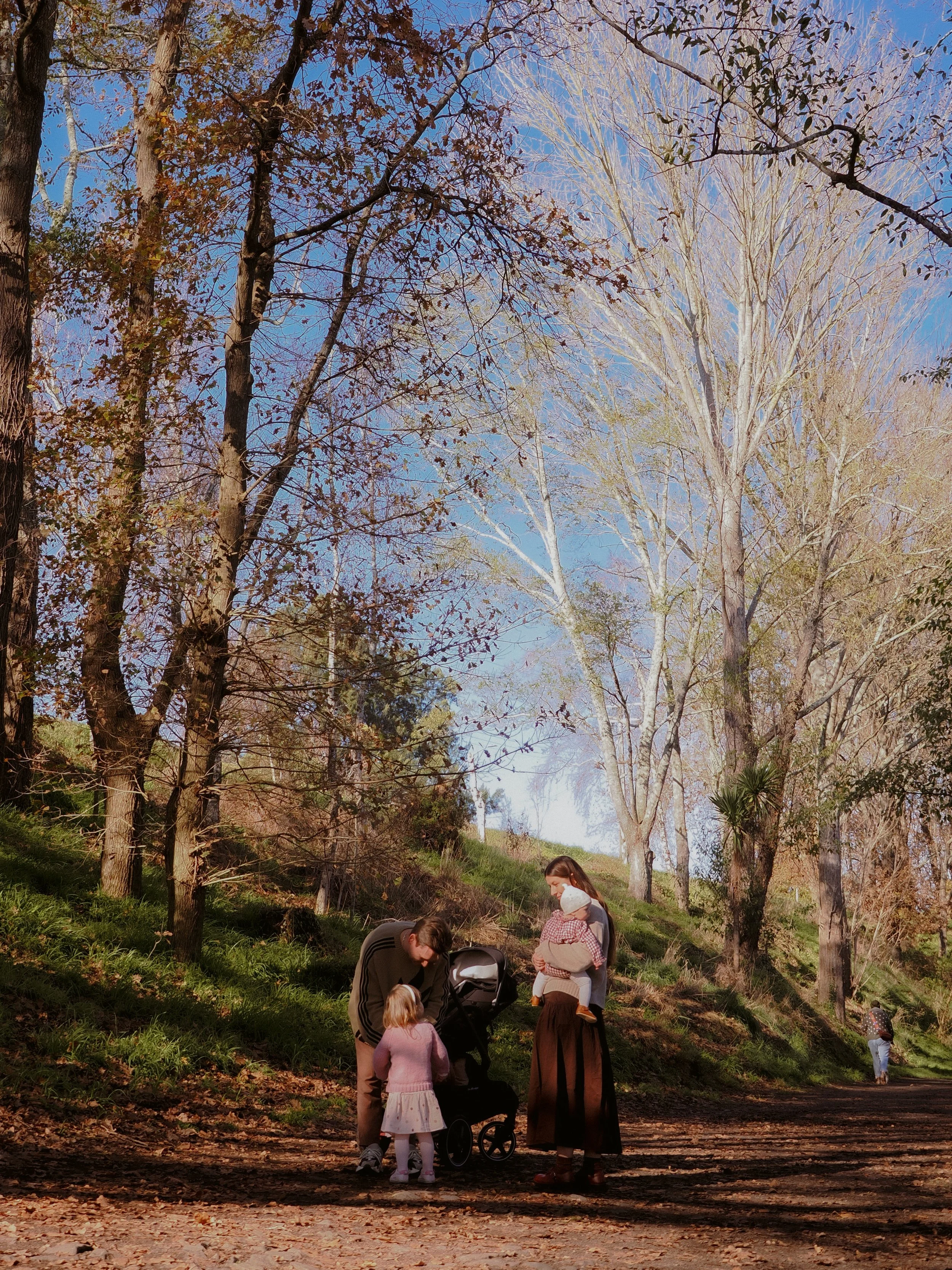 Family with a mother, father, and two young children standing on a dirt path in a wooded area during fall, with trees and a clear blue sky in the background. Family Photography Waikato New Zealand. Family Photography Hamilton New Zealand. 