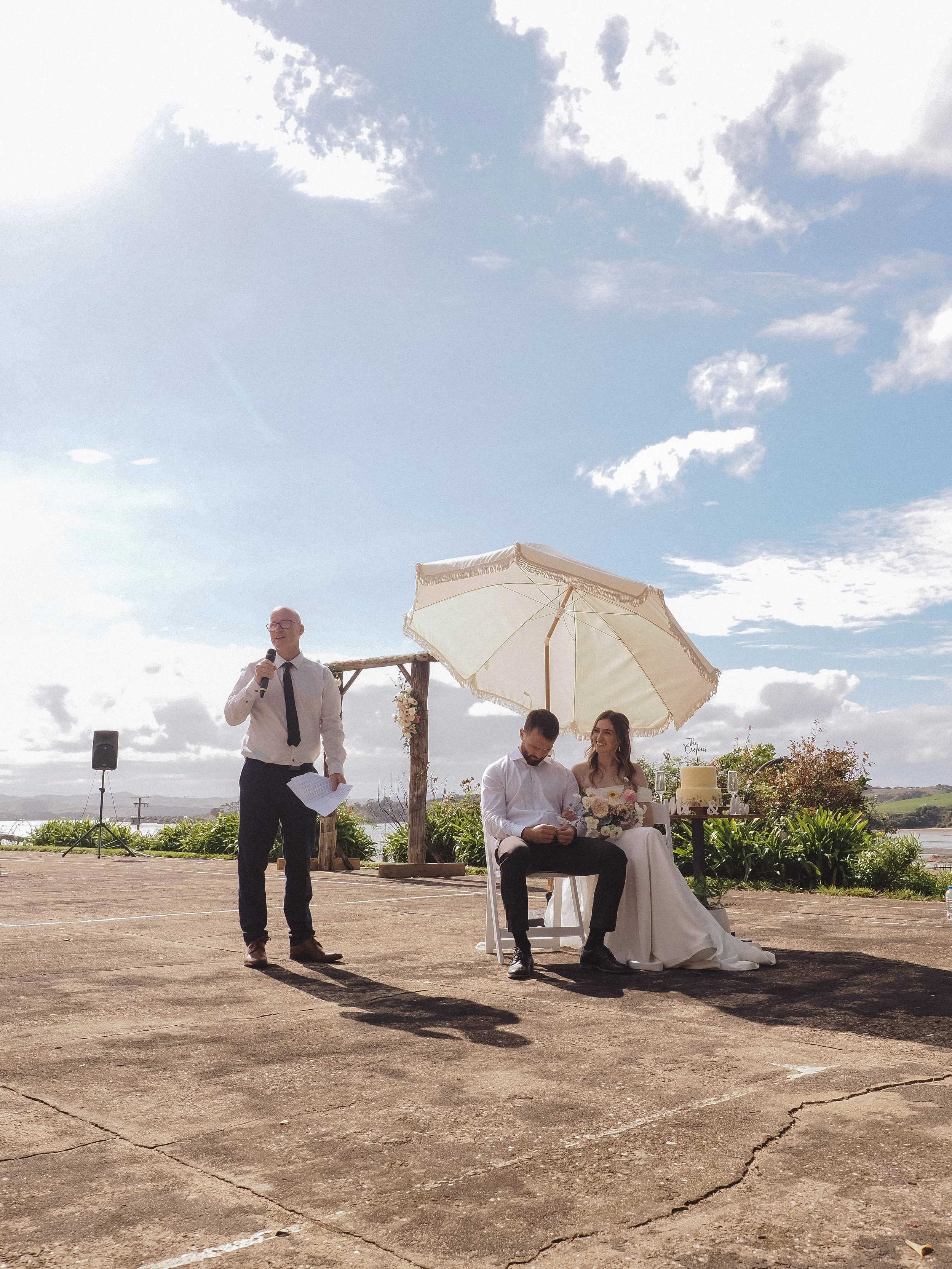 A wedding ceremony outdoors with a bride and groom seated under an umbrella, a man standing and speaking into a microphone, and a wedding cake on a table nearby.  Wedding Photography Waikato New Zealand. Wedding Photography Hamilton New Zealand. 