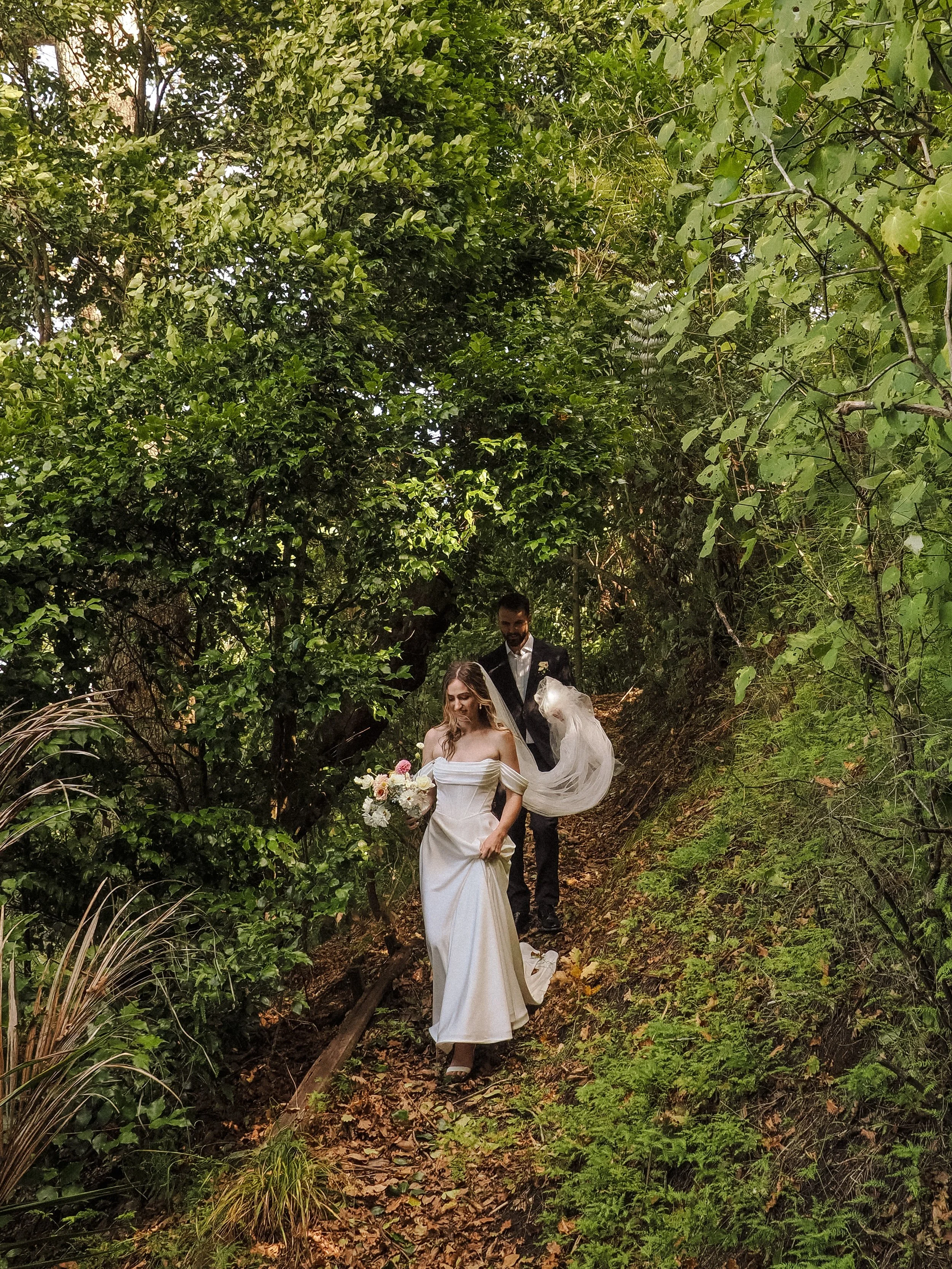 Bride and groom walking down a wooded trail; the bride is in a white gown holding a bouquet, and the groom is in a dark suit.  Wedding Photography Waikato New Zealand. Wedding Photography Hamilton New Zealand. Northland Wedding Photography