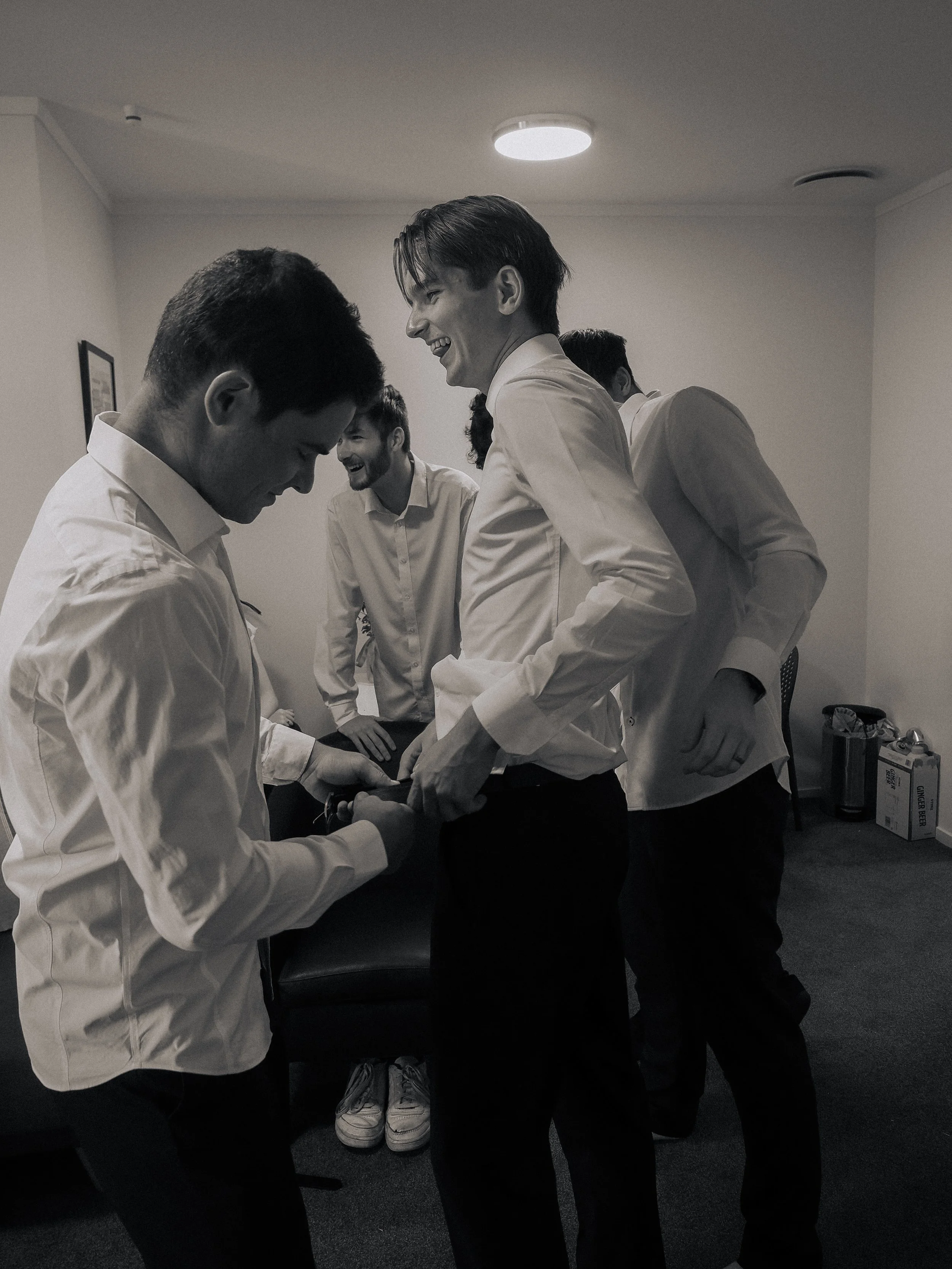 Group of young men in formal attire laughing and smiling in an indoor room, with some engaging in conversation.