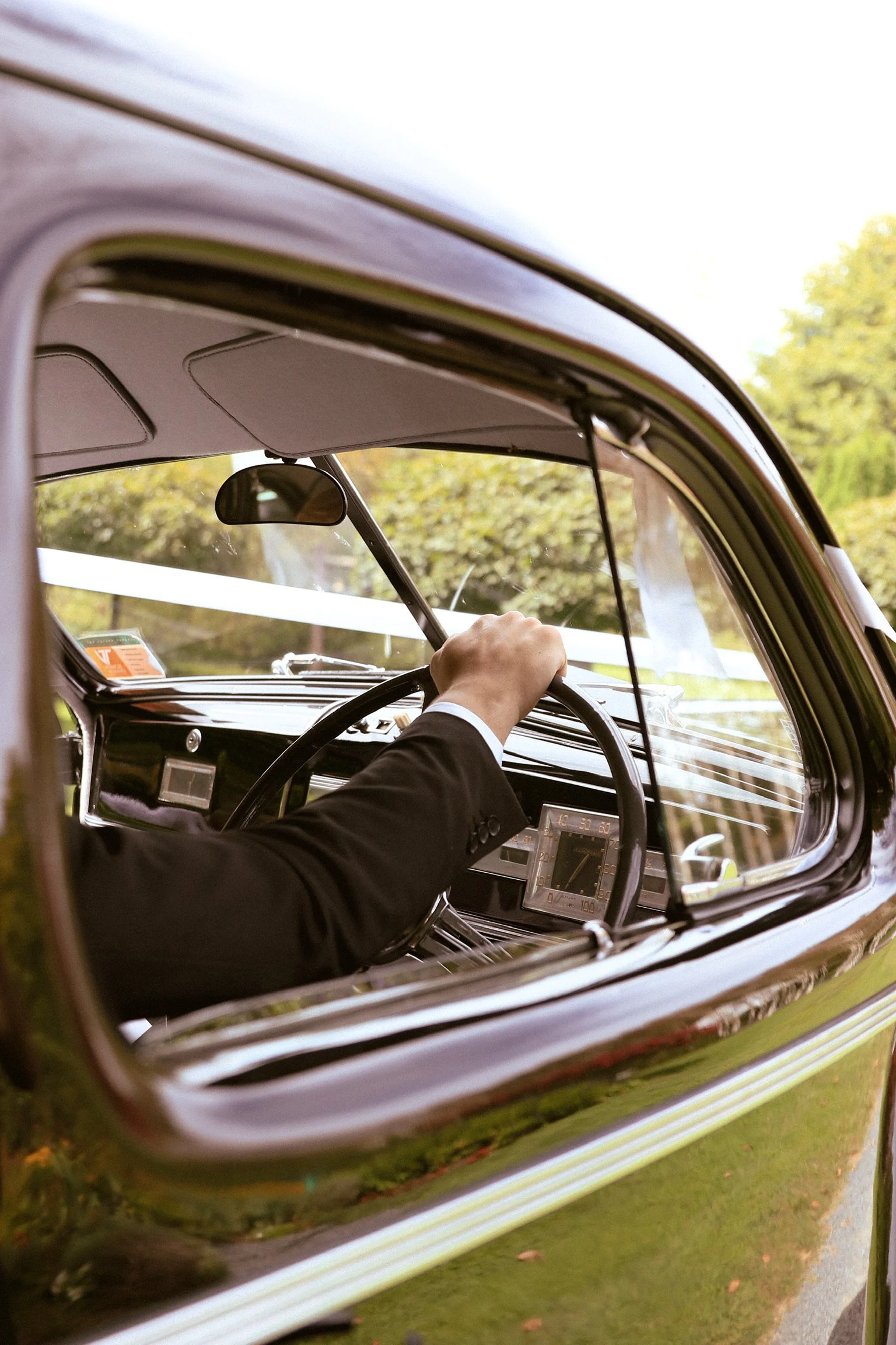Close-up view of a person driving a vintage car, with a focus on their arm and hand on the steering wheel, seen through the driver's side window. Wedding Photography Waikato New Zealand. Wedding Photography Hamilton New Zealand