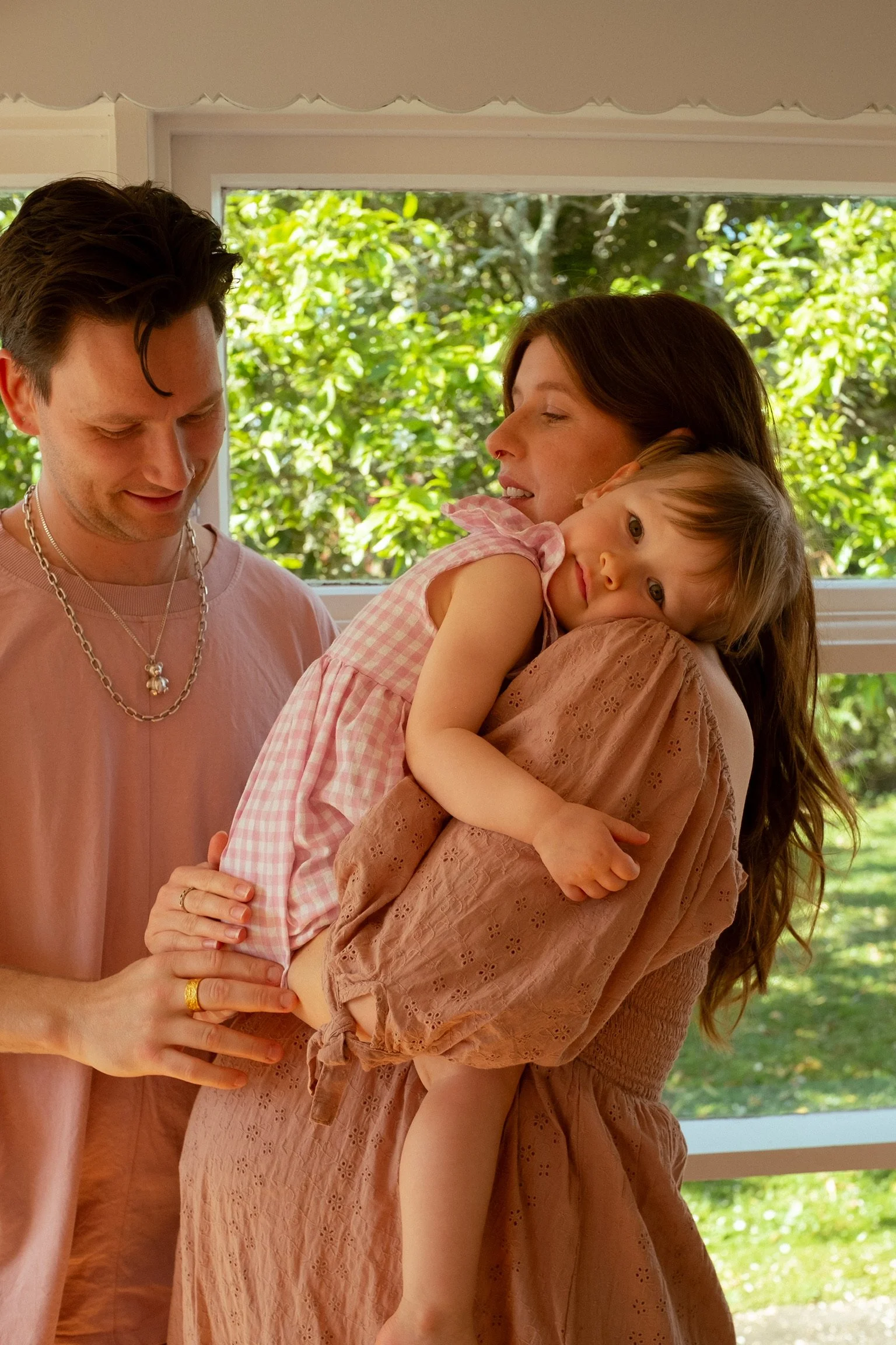 A woman holds a young girl, who rests her head on her shoulder, while a man stands beside them, all smiling softly. Maternity Photography. Family Photographer. Hamilton New Zealand Photographer