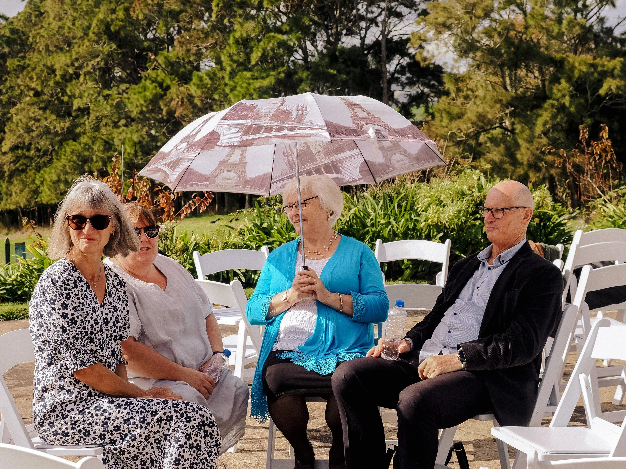 Four people sitting outdoors, with two women wearing sunglasses, one woman holding an umbrella with a Paris Eiffel Tower print, and a man in a suit with glasses, surrounded by white chairs and lush greenery in the background.