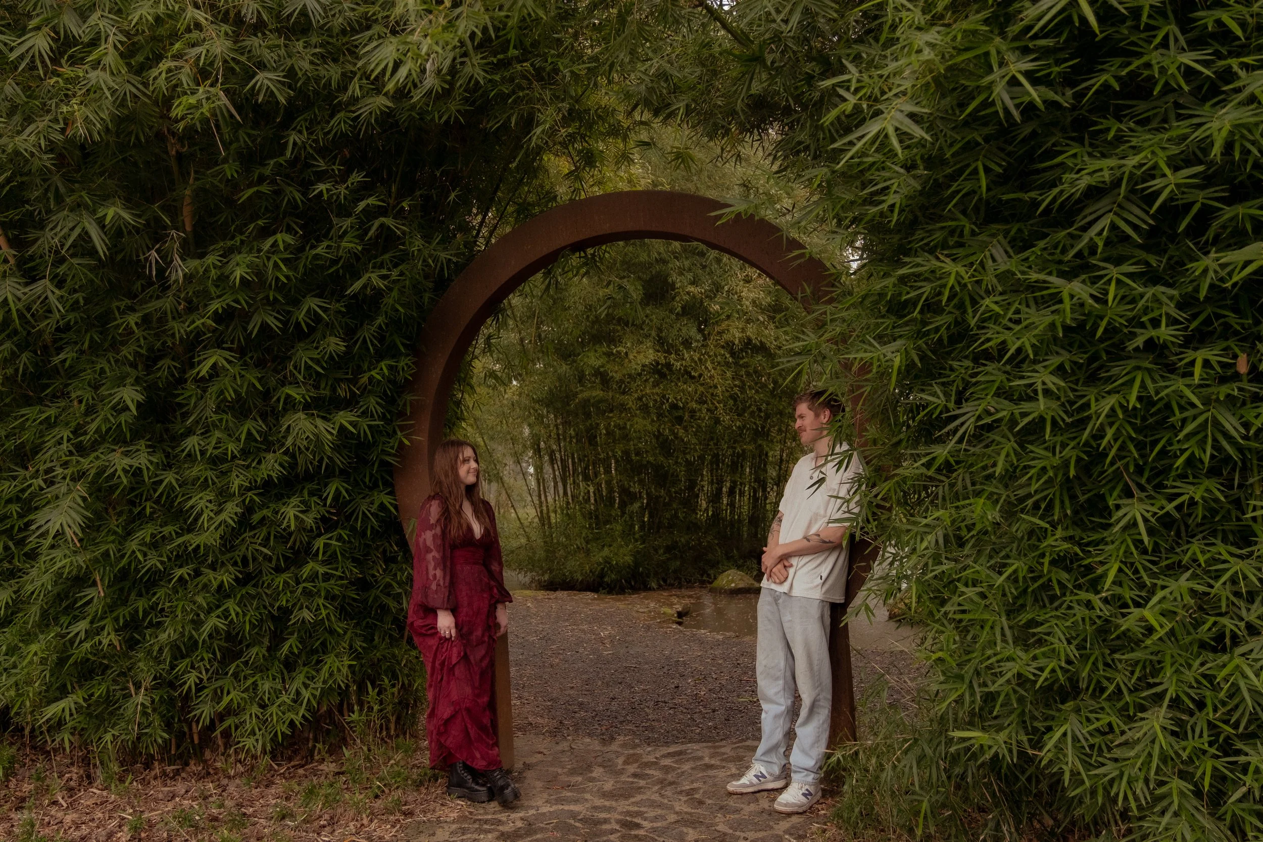 Two people standing on a dirt path beside a circular metal archway surrounded by dense green foliage, with trees and water in the background. Couples photography Raglan New Zealand. Engagement photography Hamilton New Zealand. 