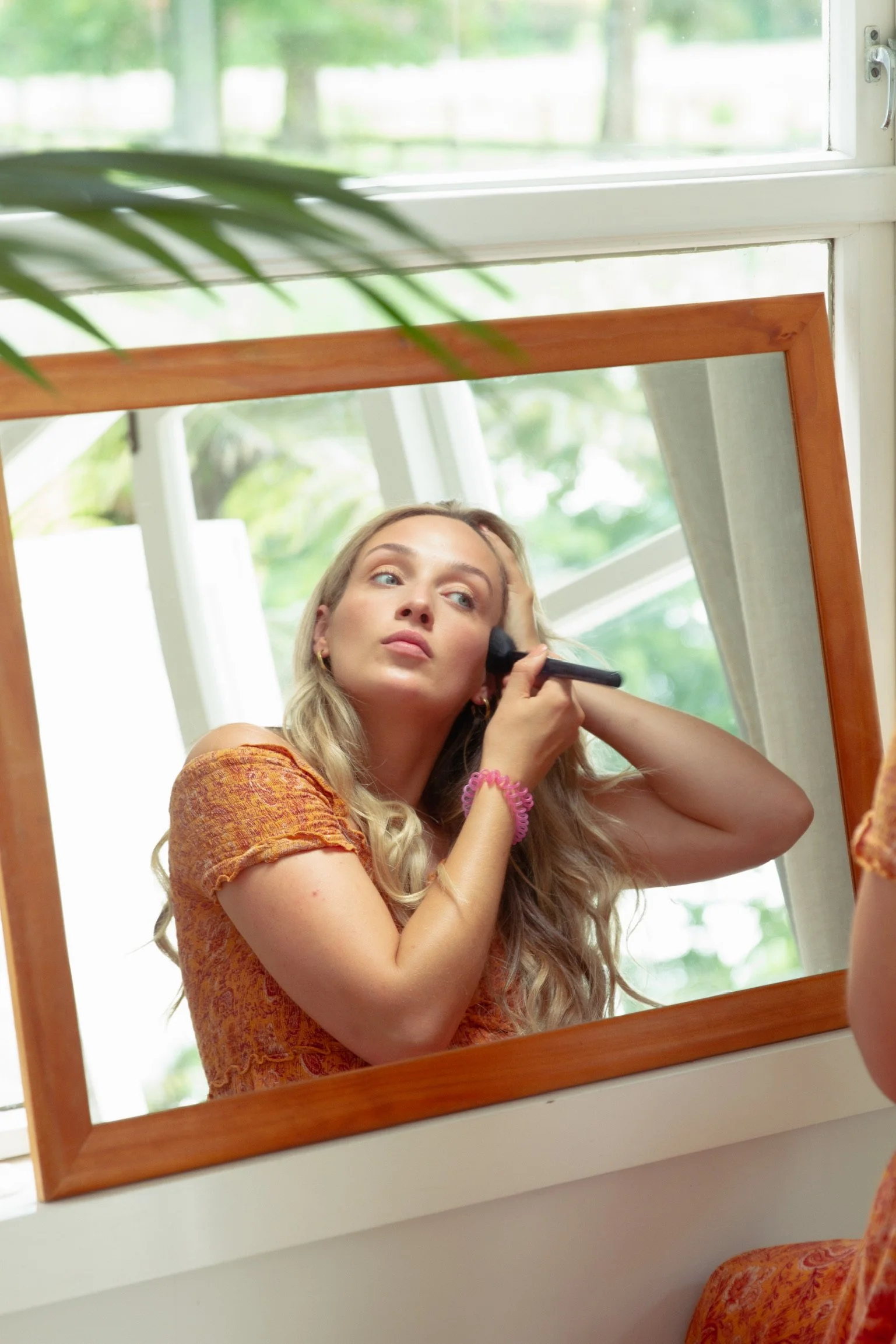 A young woman with blonde hair in a loose braid, wearing an orange dress, is applying makeup using a brush while looking into a mirror. She is sitting in front of a window with a wooden frame, and greenery is visible outside.
