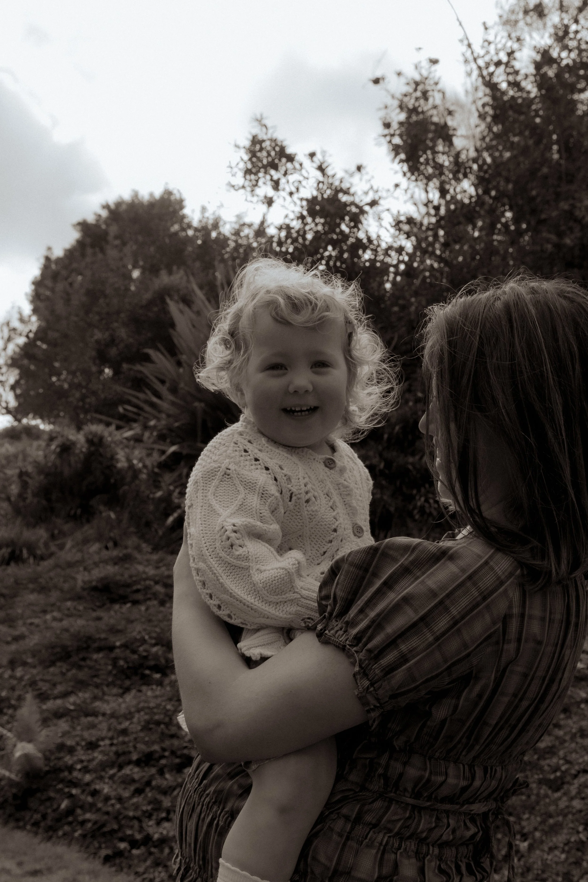 A woman holding a smiling young girl outdoors with trees and cloudy sky in the background. Family photographer, maternity photography, Hamilton New Zealand photographer