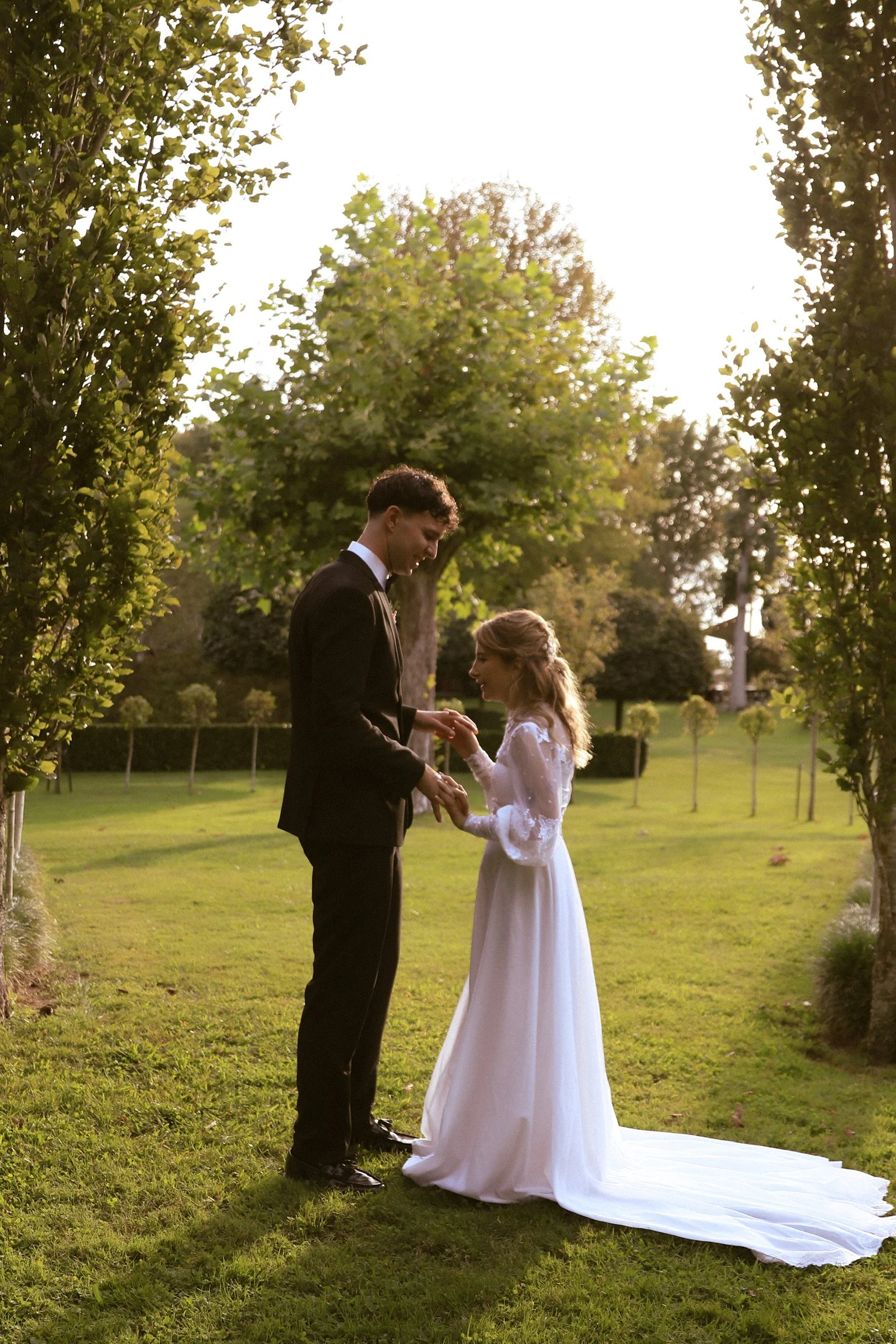 A couple dressed in wedding attire standing in a garden, holding hands and exchanging rings during sunset.