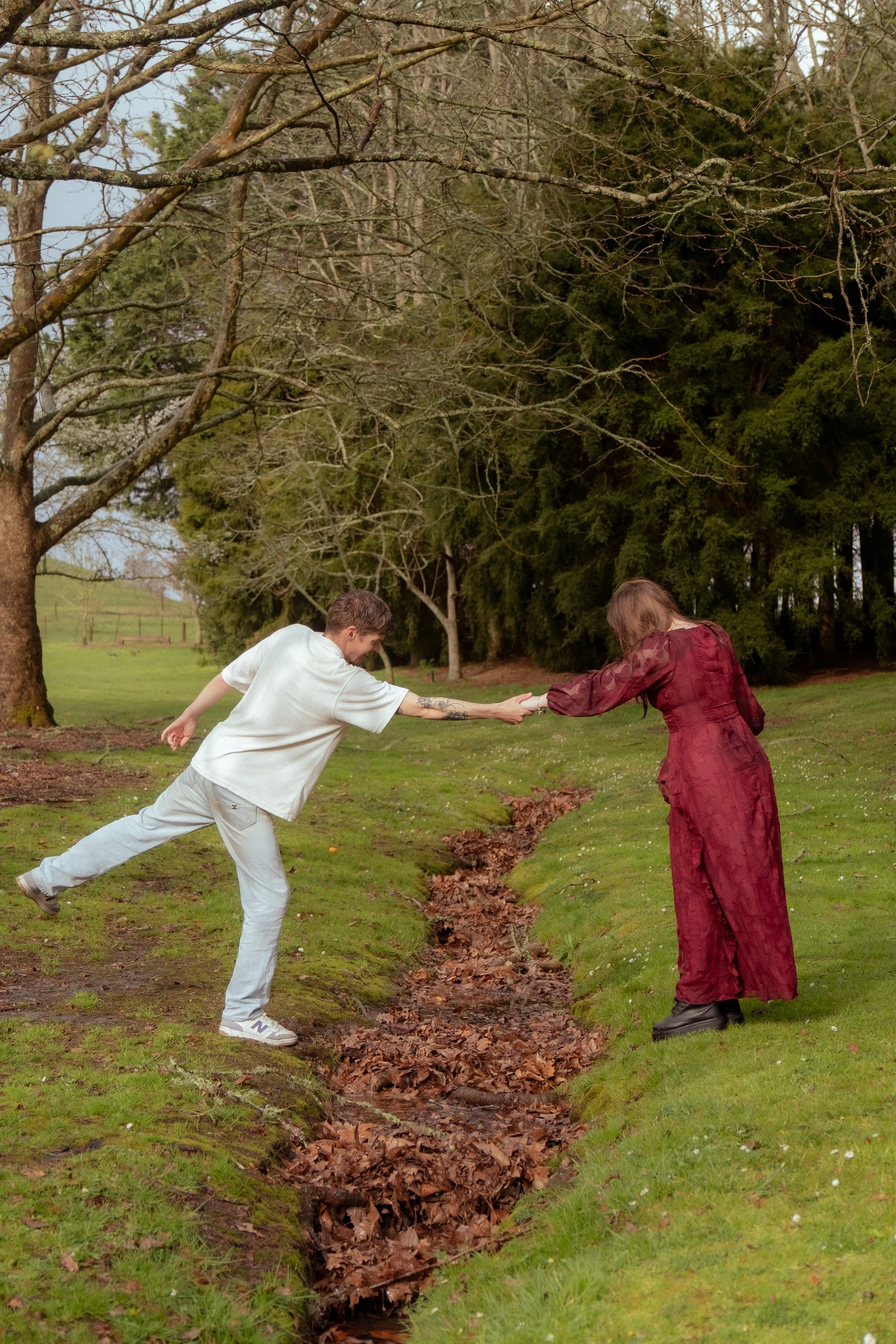 Two people shake hands over a small creek in a park with trees in the background. Couples photography Hamilton New Zealand. Engagement photography Hamilton New Zealand. Whangarei New Zealand Couples photographer