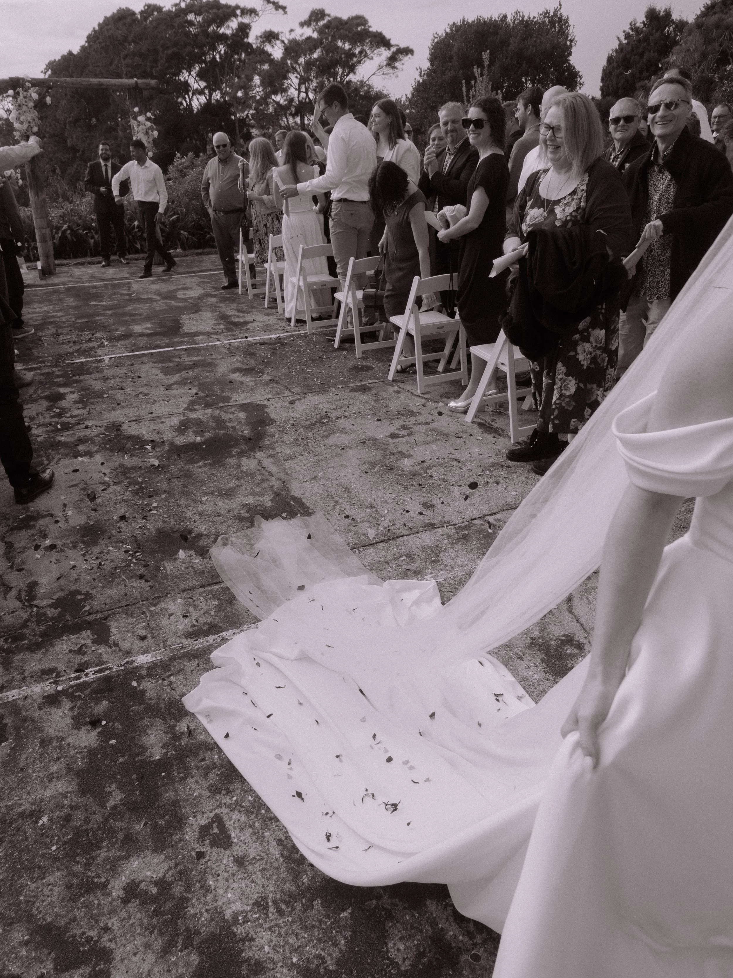 A bride in a white wedding gown is standing outdoors on a concrete surface, with her gown train on the ground. People, including men and women, are standing and sitting on white chairs, smiling and watching a wedding ceremony. Some are dressed formal