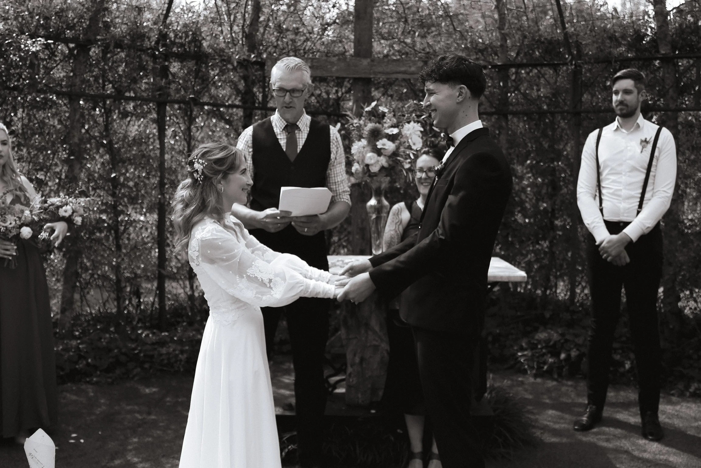 A black and white photo of a wedding ceremony outdoors, with a bride and groom holding hands and exchanging vows, surrounded by four guests watching, with trees and a fence in the background. Wedding Photography Waikato New Zealand. 