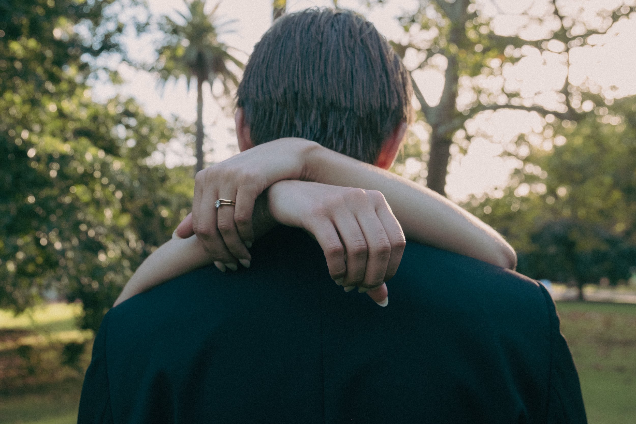 A couple embracing outdoors with the woman’s face hidden and arms around each other, trees and sunlight in the background.