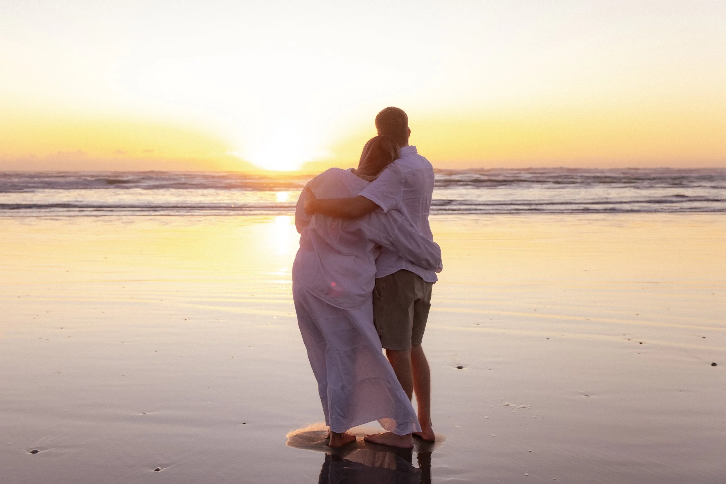A couple embracing and watching the sunset on a beach. Maternity photography. Raglan New Zealand Photographer