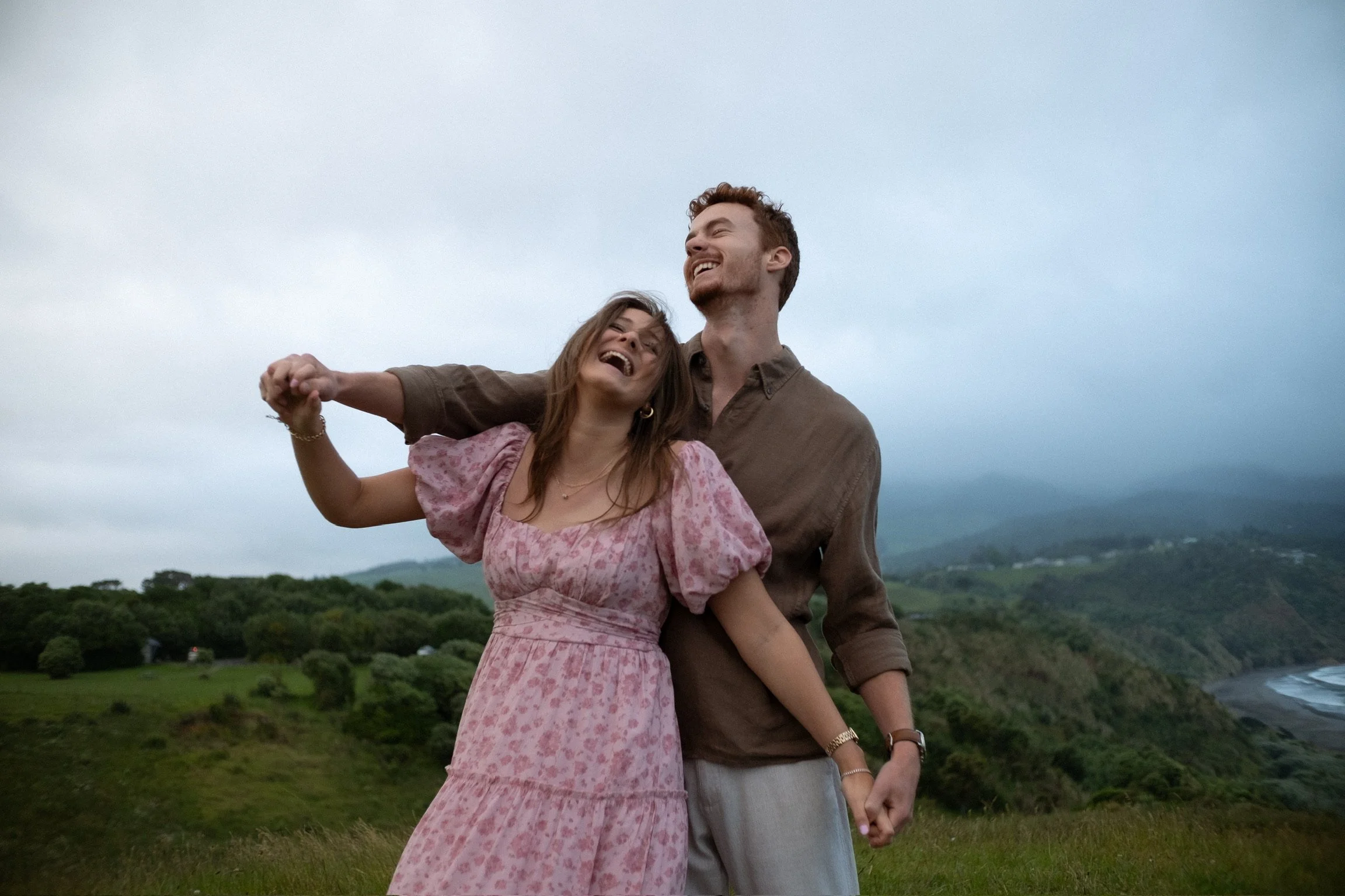 A couple dancing and laughing outdoors in a green hilly landscape with a cloudy sky. Couples photography Raglan New Zealand. Engagement photography Hamilton New Zealand. Whangarei New Zealand Couples photographer