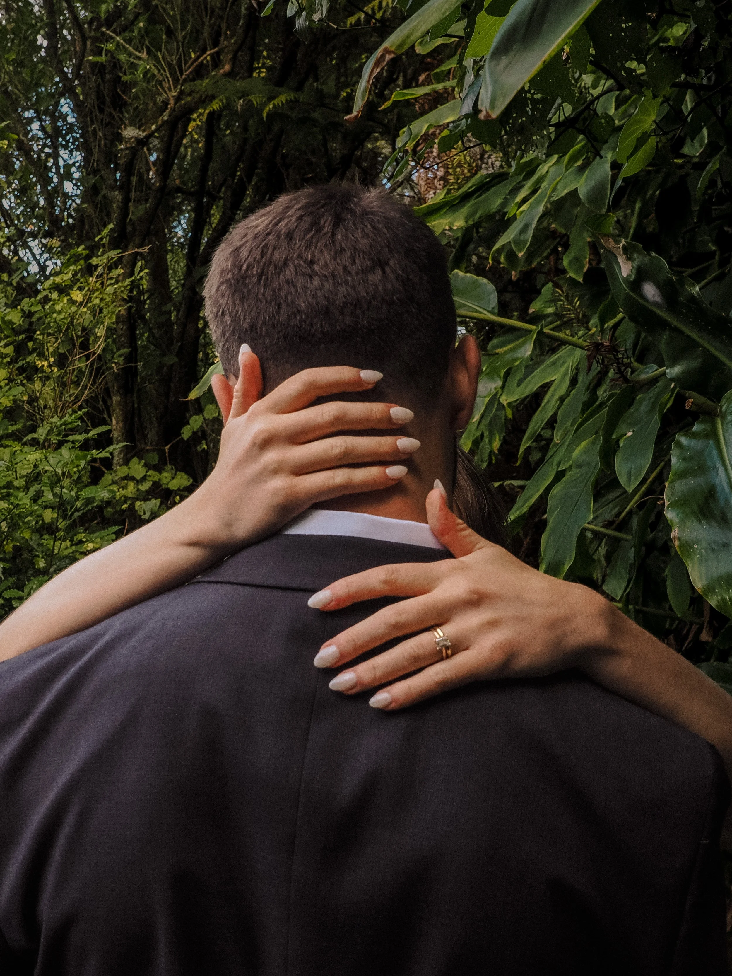 Person with short hair in a suit embracing another person, with their hand gently touching the back of the other's head, in a lush green outdoor setting.  Wedding Photography Waikato New Zealand. Wedding Photography Hamilton New Zealand. Northland 