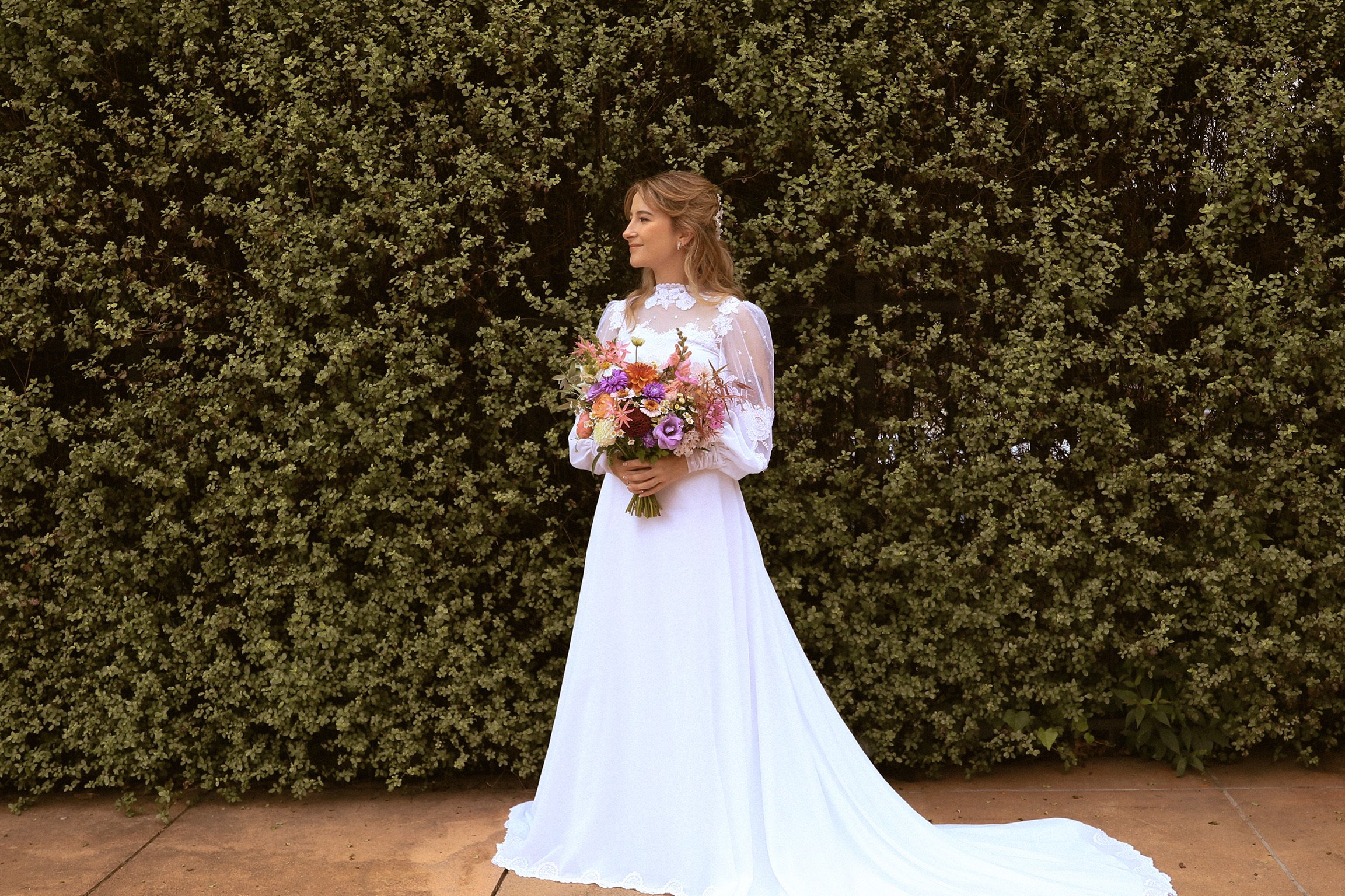A bride in a white wedding gown holding a colorful bouquet of flowers standing outdoors in front of green foliage. Wedding Photography Waikato New Zealand. Wedding Photography Hamilton New Zealand