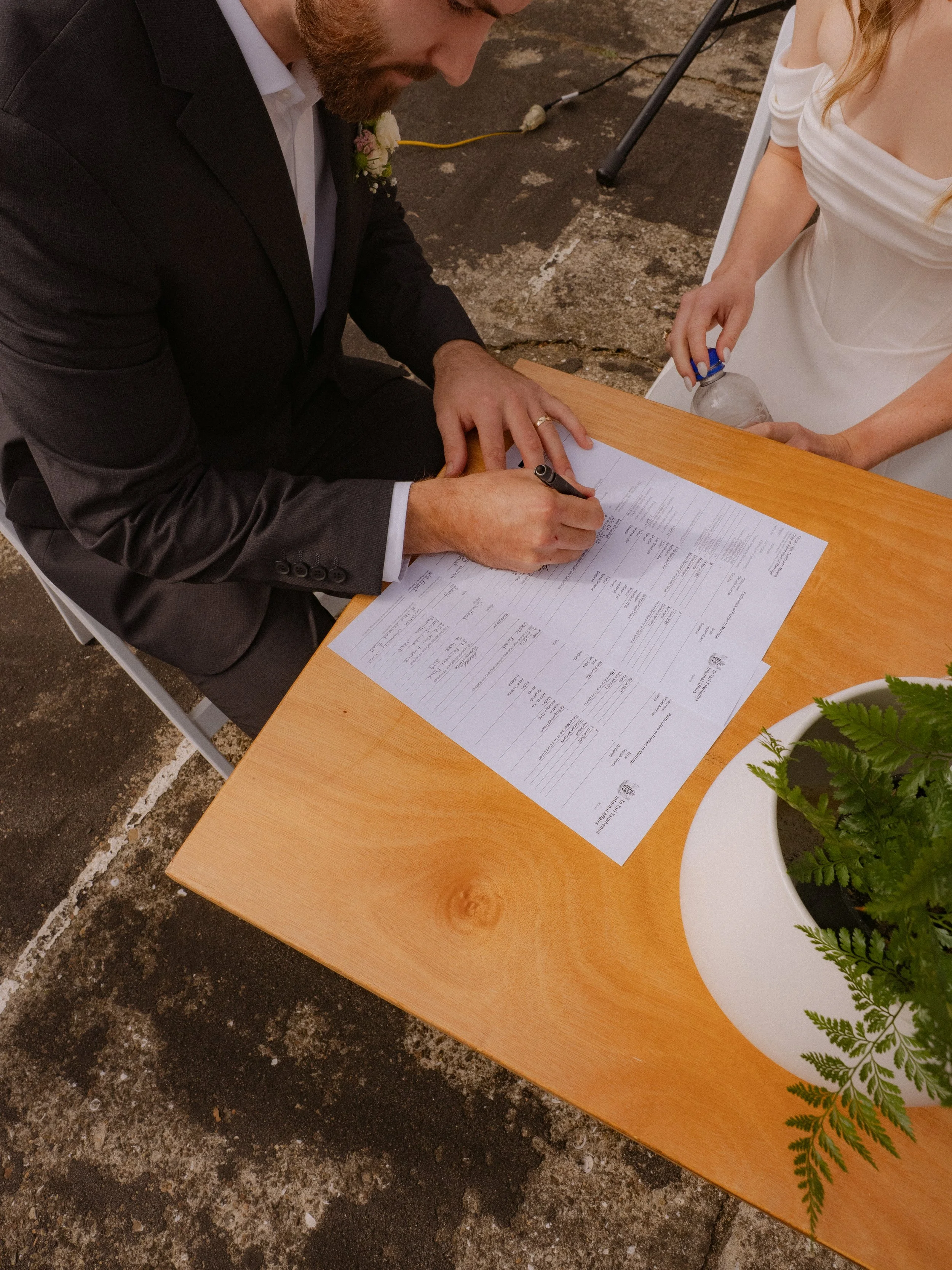 A man in a black suit with a boutonniere signs a document on a wooden table, while a woman in a white dress holds a water bottle nearby.  Wedding Photography Waikato New Zealand. Wedding Photography Hamilton New Zealand. Northland Wedding Photography