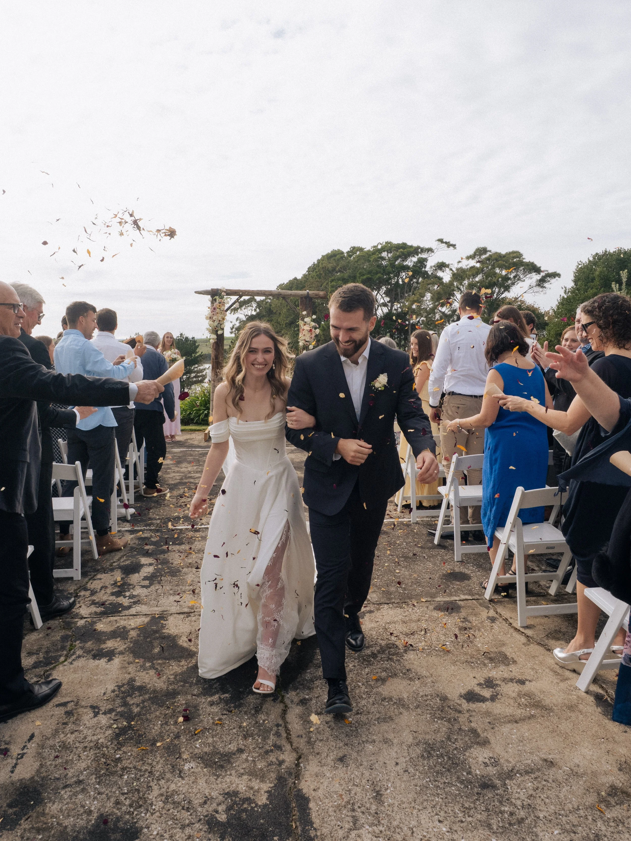 A newlywed couple walks down the aisle at an outdoor wedding, smiling and surrounded by guests throwing confetti.  Wedding Photography Waikato New Zealand. Wedding Photography Hamilton New Zealand. Northland Wedding Photography
