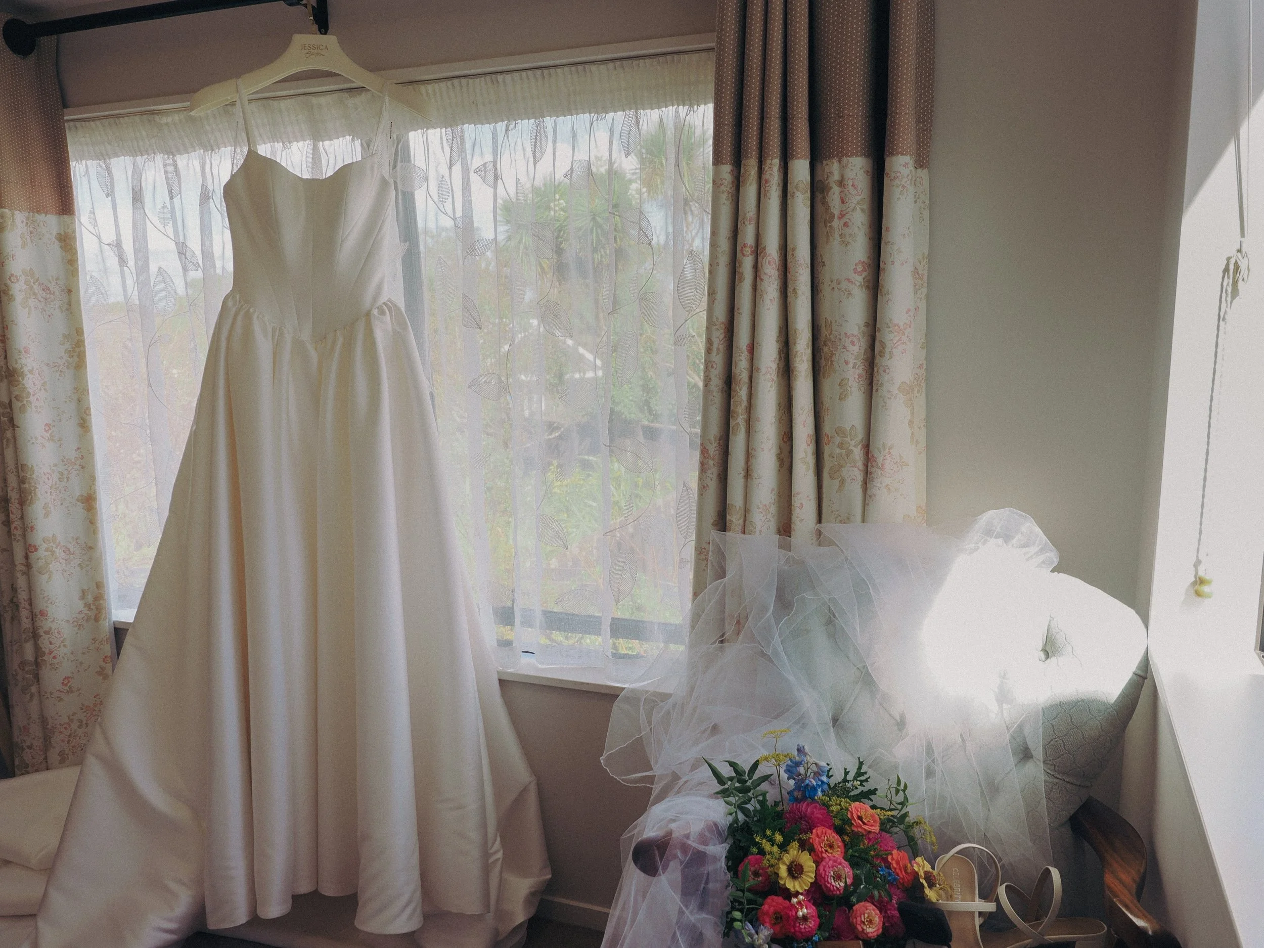 A white dress hanging on a hook in front of a window with floral curtains, next to a bouquet of colorful flowers wrapped in tulle.