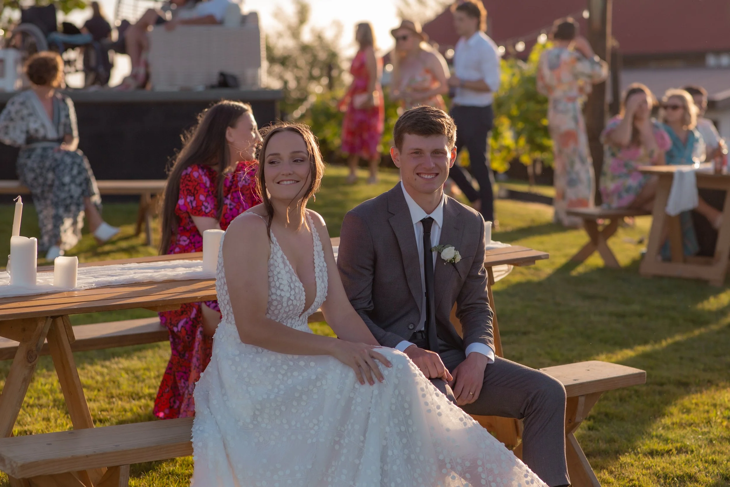 A bride and groom sitting on a wooden bench at their outdoor wedding reception during sunset, smiling and holding hands, surrounded by guests in floral attire.  Wedding Photography Waikato New Zealand. Wedding Photography Hamilton New Zealand. 