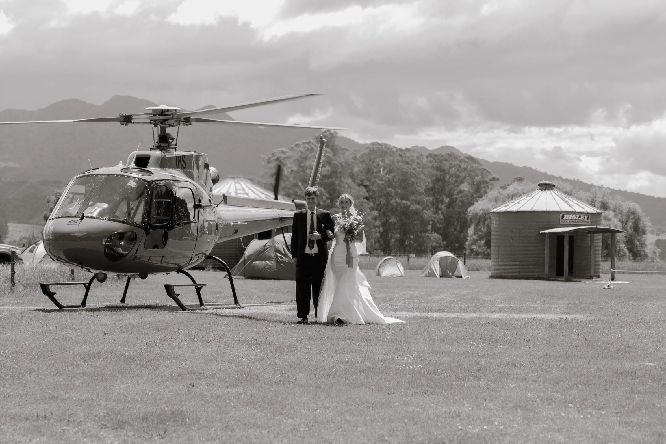 A wedding couple walking hand in hand near a helicopter on a grassy field with tents and one round building in the background, surrounded by mountains and cloudy sky.  Wedding Photography Waikato New Zealand. Wedding Photography Hamilton New Zealand.