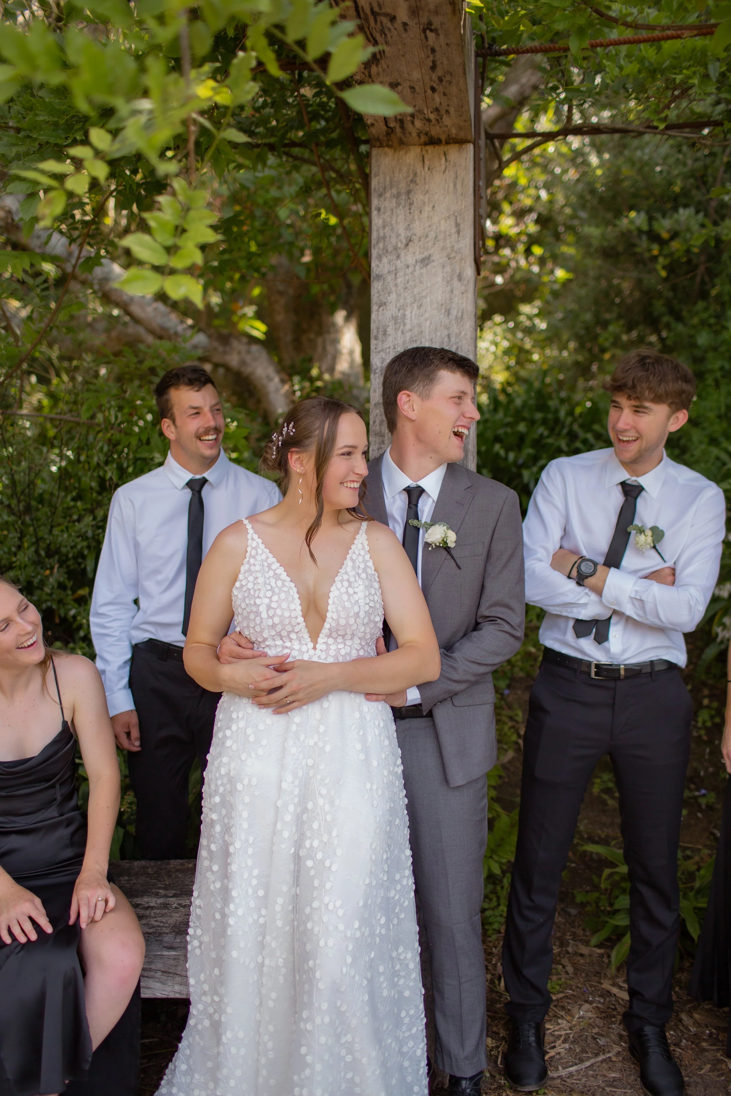 A group of wedding guests, including a bride in a white dress and a groom in a gray suit, laughing and smiling outdoors among green trees and foliage.  Wedding Photography Waikato New Zealand. Wedding Photography Hamilton New Zealand. Northland 