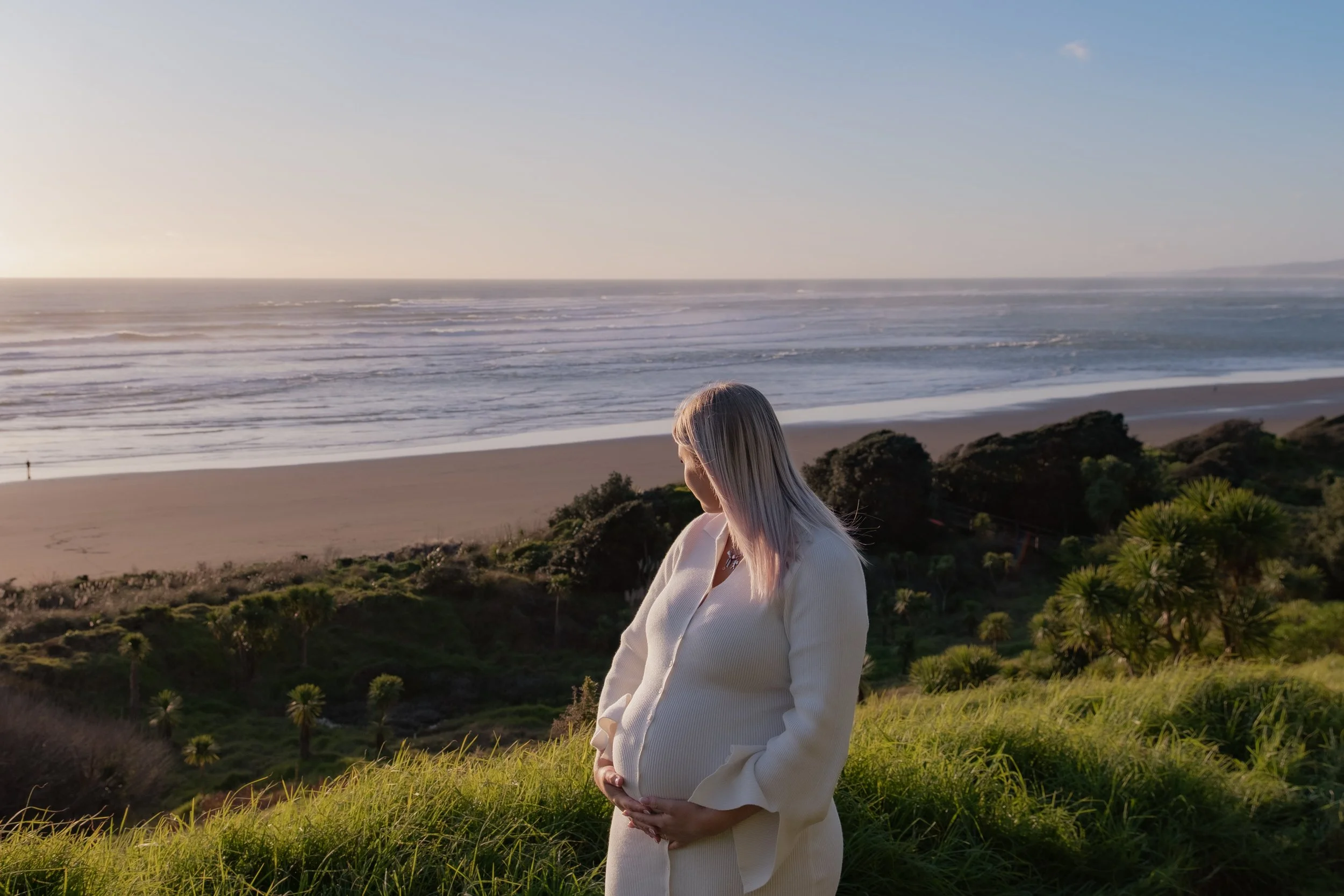 A pregnant woman with light-colored hair, wearing a cream-colored sweater and pants, stands on a grassy hill overlooking a sandy beach and ocean during sunset, with a person walking along the shoreline in the background. Raglan New Zealand