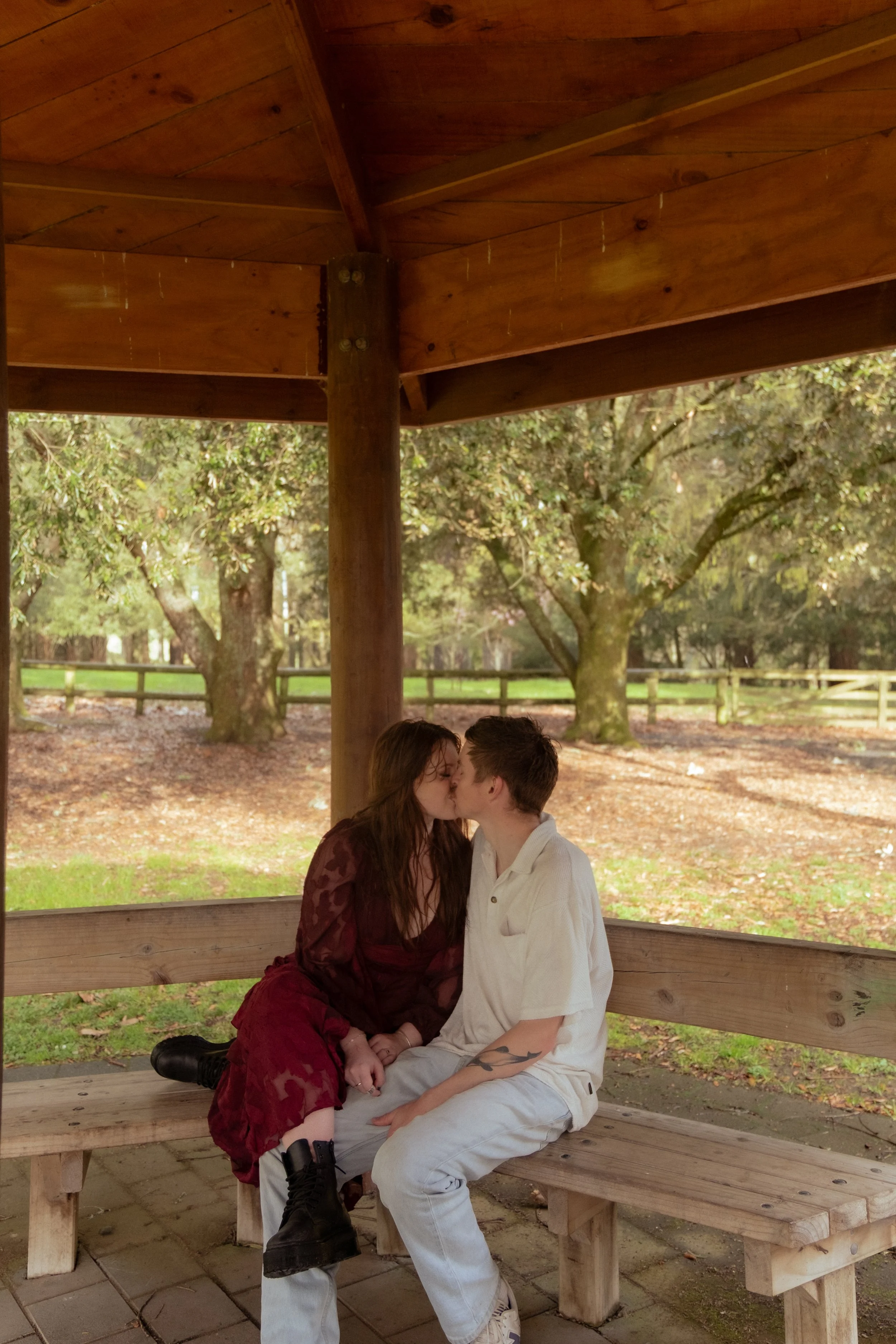 A couple sitting on a wooden bench under a gazebo, kissing outdoors in a park with trees and green grass. Couples photography Hamilton New Zealand. Engagement photography Hamilton New Zealand. Whangarei New Zealand Couples photographer