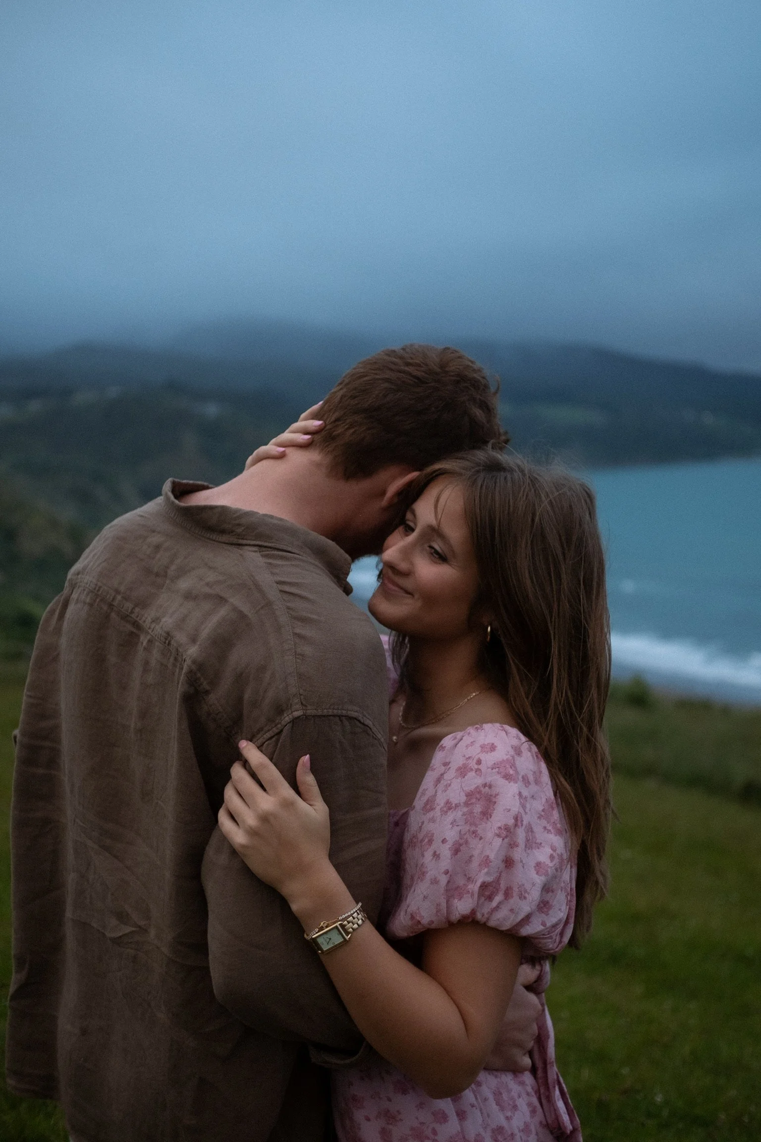 A couple embracing outdoors near the ocean, with mountains in the background, during dusk. Couples photography Raglan New Zealand. Engagement photography Hamilton New Zealand. Whangarei New Zealand Couples photographer