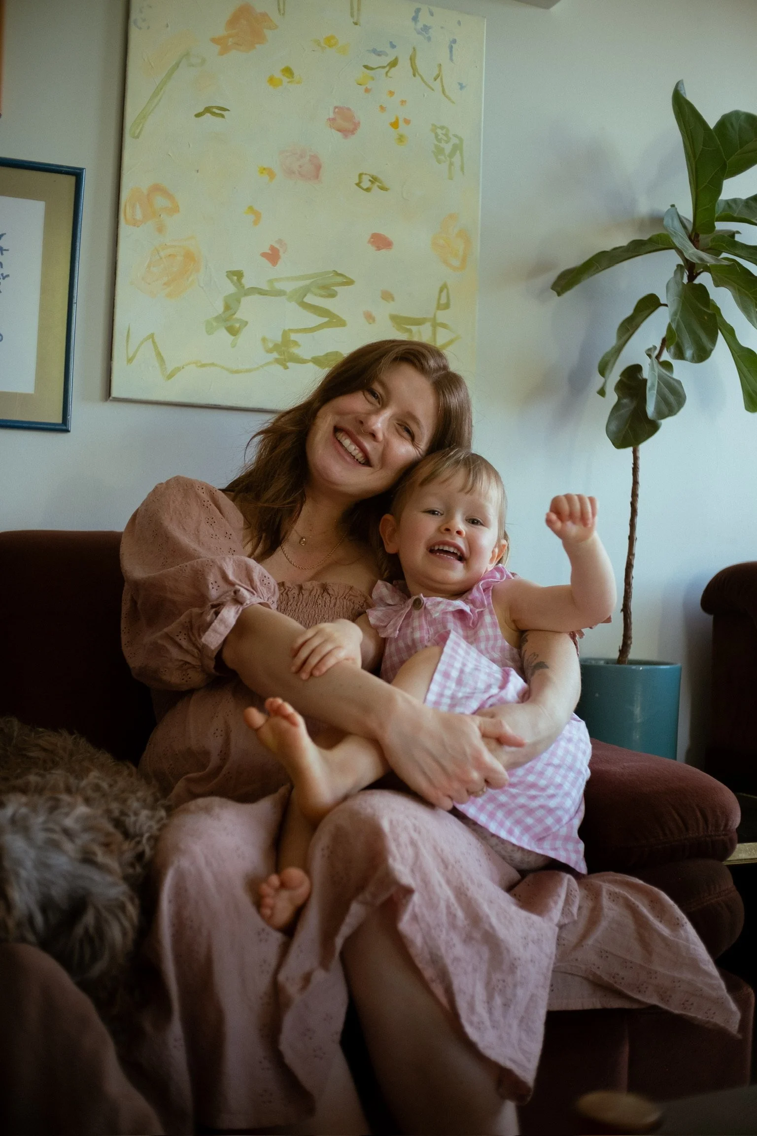 A woman and a young girl are sitting together on a couch, smiling and laughing. The woman has shoulder-length brown hair and is wearing a pink dress with puffed sleeves. The girl is wearing a pink gingham dress and has short blonde hair. They are in 