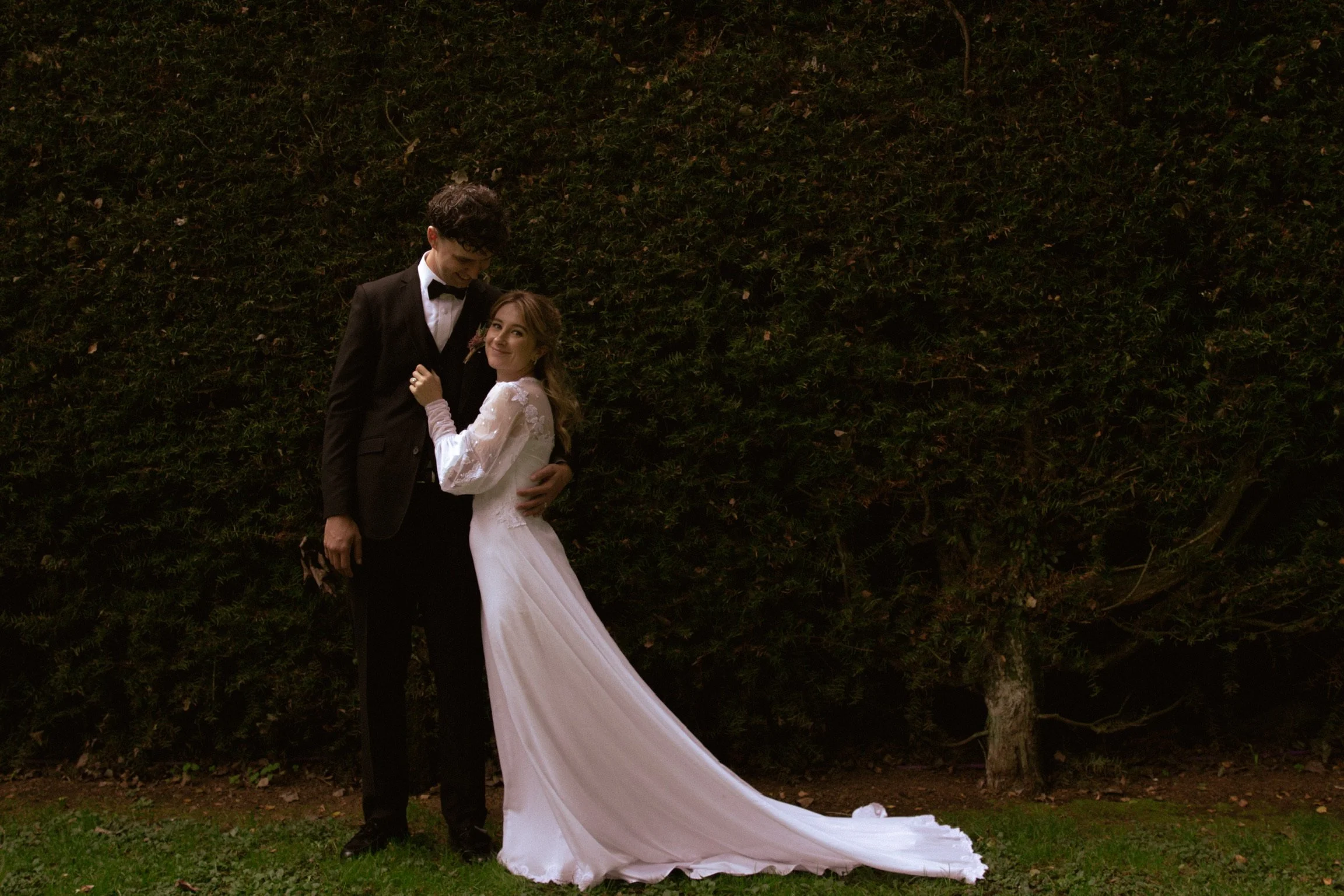 A couple in wedding attire standing outdoors at night, the groom in a black tuxedo and the bride in a white wedding dress, their arms around each other, smiling. Wedding Photography Waikato New Zealand. Wedding Photography Hamilton New Zealand