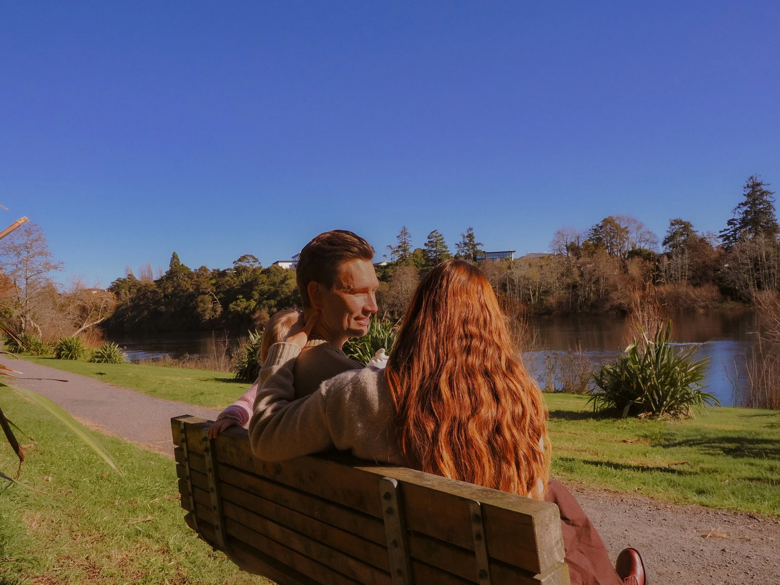 Two people sitting on a park bench by a river, enjoying a sunny day with a clear blue sky and trees in the background. Family Photography Waikato New Zealand. Family Photography Hamilton New Zealand. Northland Wedding Photography