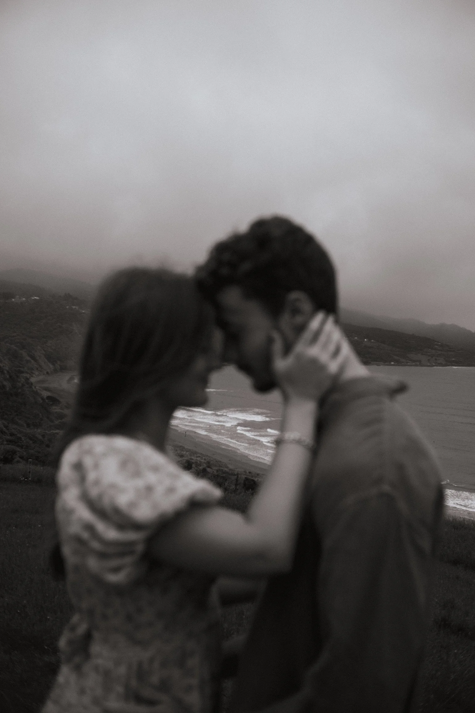 A black-and-white photo of a couple holding each other close near a beach, their faces touching gently, with ocean waves and hills in the background. Couples Photography Raglan New Zealand. Documentary photographer Whanagrei New Zealand