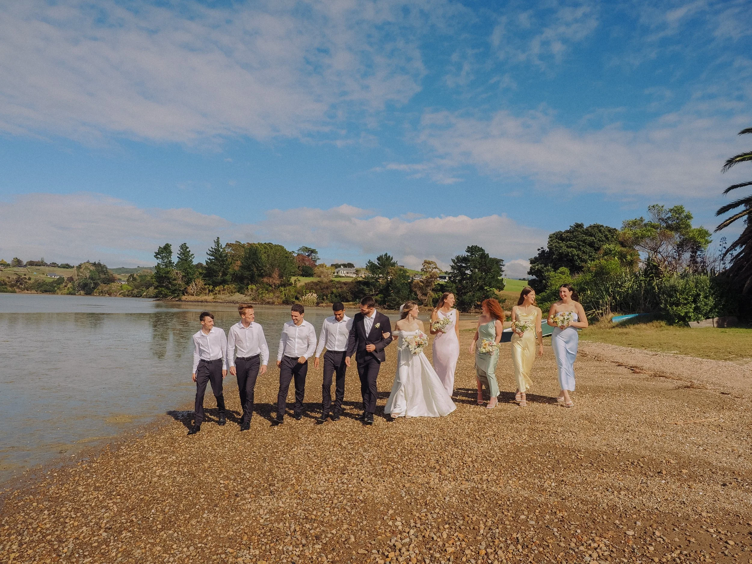Group of people in formal attire walking on a pebbled shore near a body of water on a clear day with blue sky and trees in the background.  Wedding Photography Waikato New Zealand. Wedding Photography Hamilton New Zealand. Northland Wedding Photo