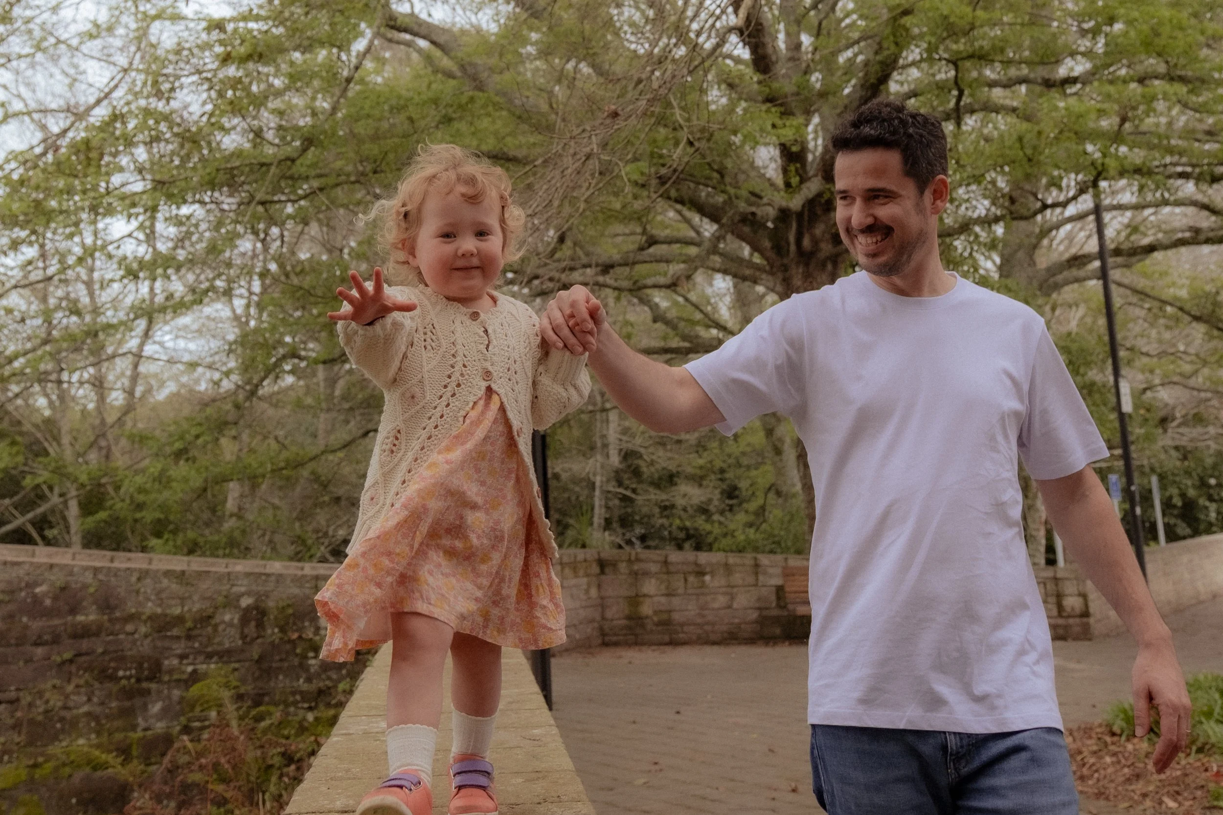 A man and a young girl hold hands and walk outdoors on a path with trees in the background. The girl appears to be balancing on a low wall, stretching her arm forward, wearing a cream cardigan and a pink dress. The man, dressed in a white t-shirt and